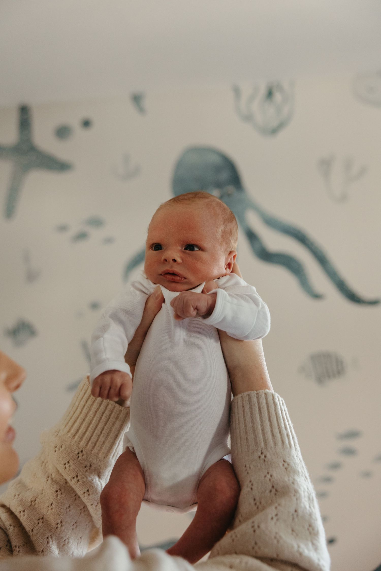 A mother holds her newborn baby up in a nursery with an ocean-themed wallpaper featuring jellyfish and starfish.