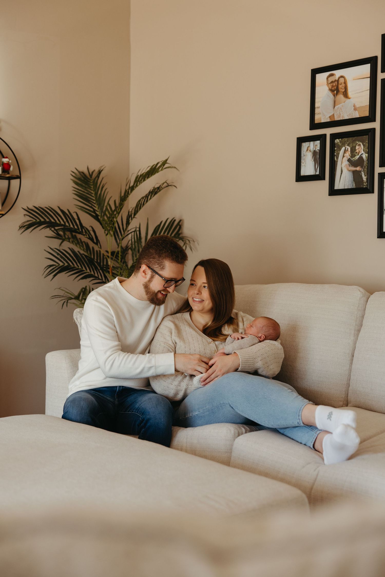 A loving couple cuddles their newborn baby on a white couch with framed photos and a plant in the background.