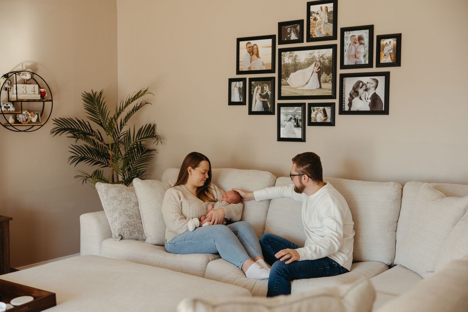 A young couple sits on a white sofa admiring their newborn baby, surrounded by framed family photos on the wall.