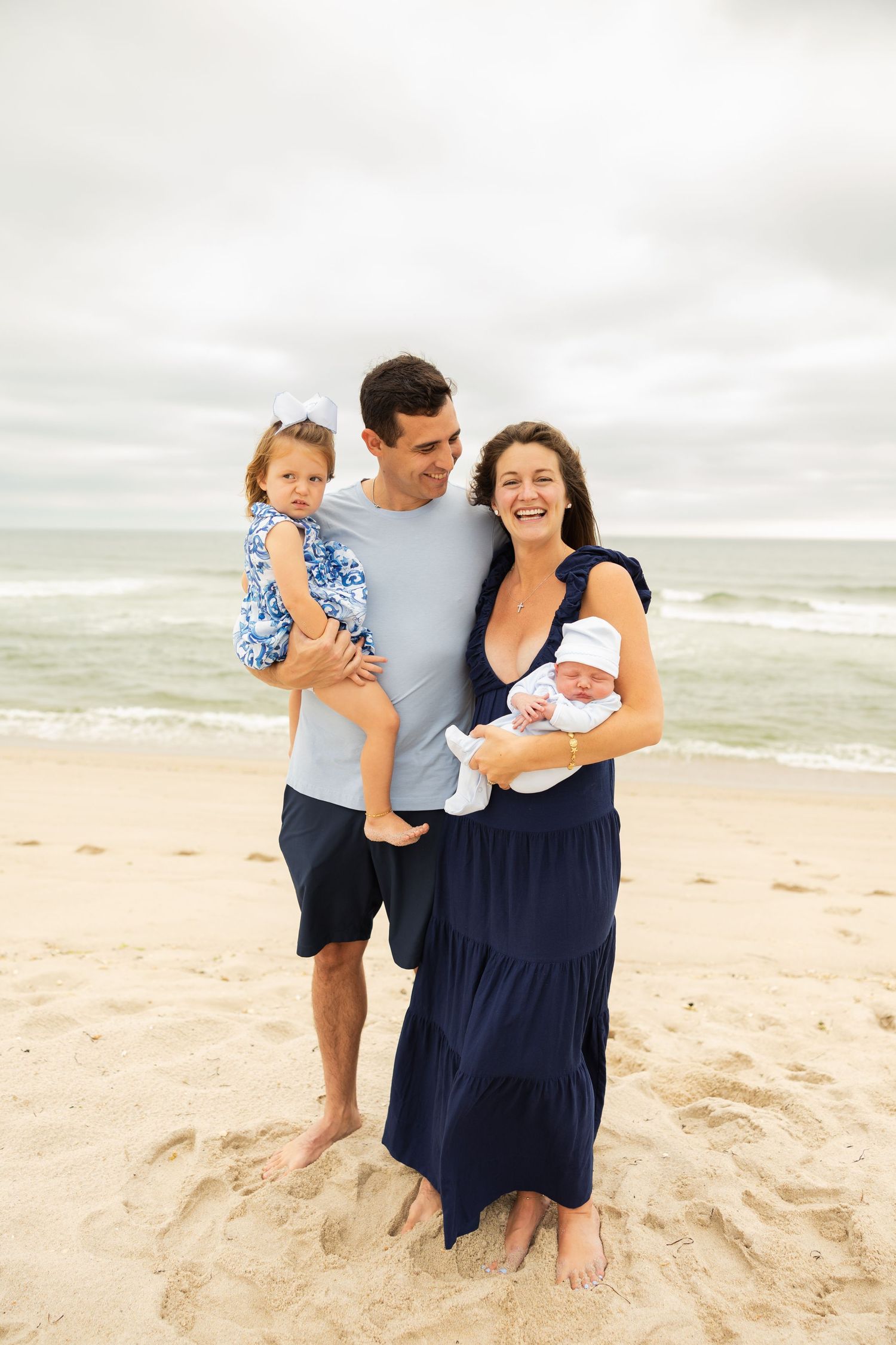 A family stands together on a sandy beach with waves and cloudy skies in the background.