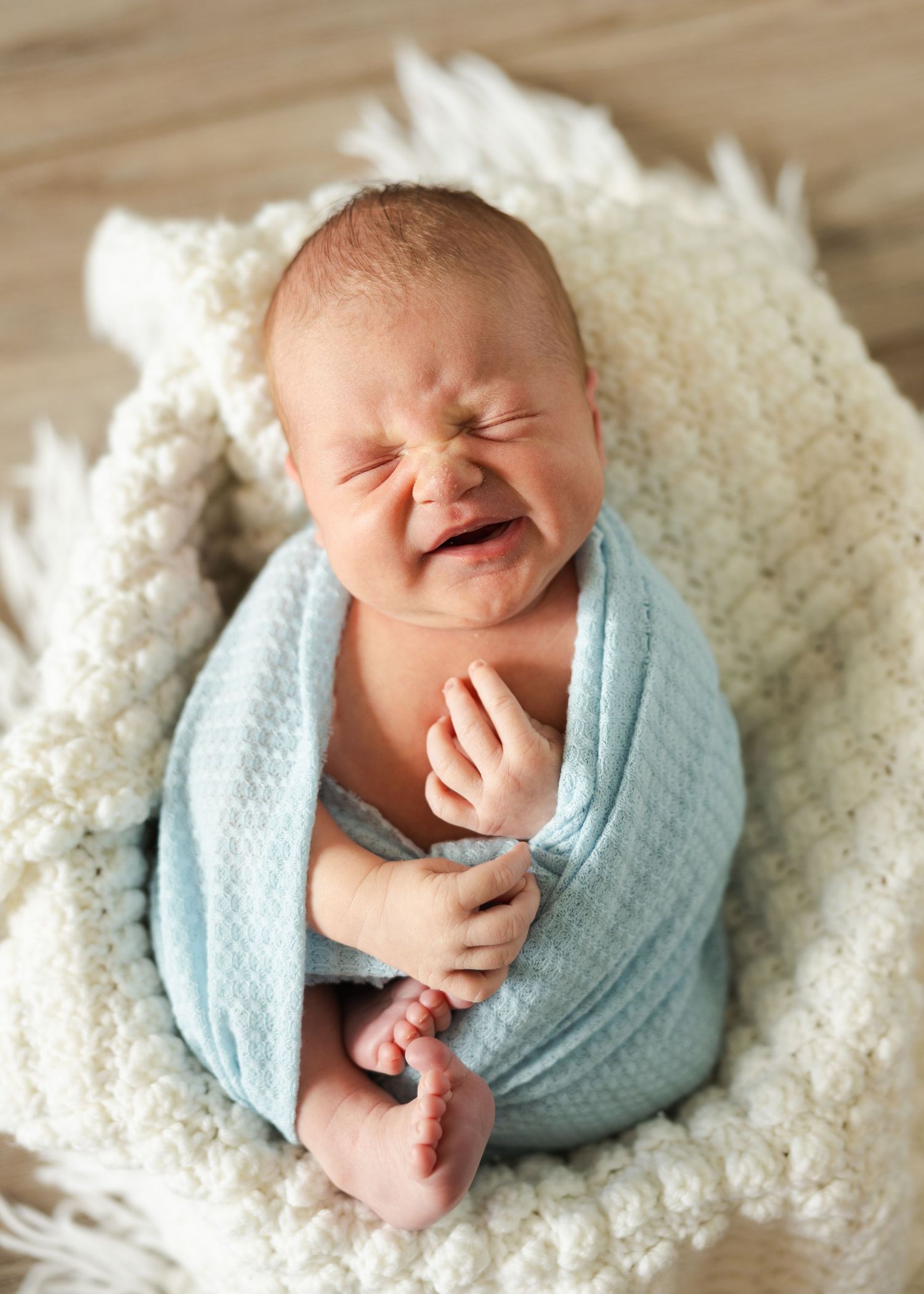 A sleeping newborn baby wrapped in a soft blue swaddle blanket lies on a cozy white textured blanket.