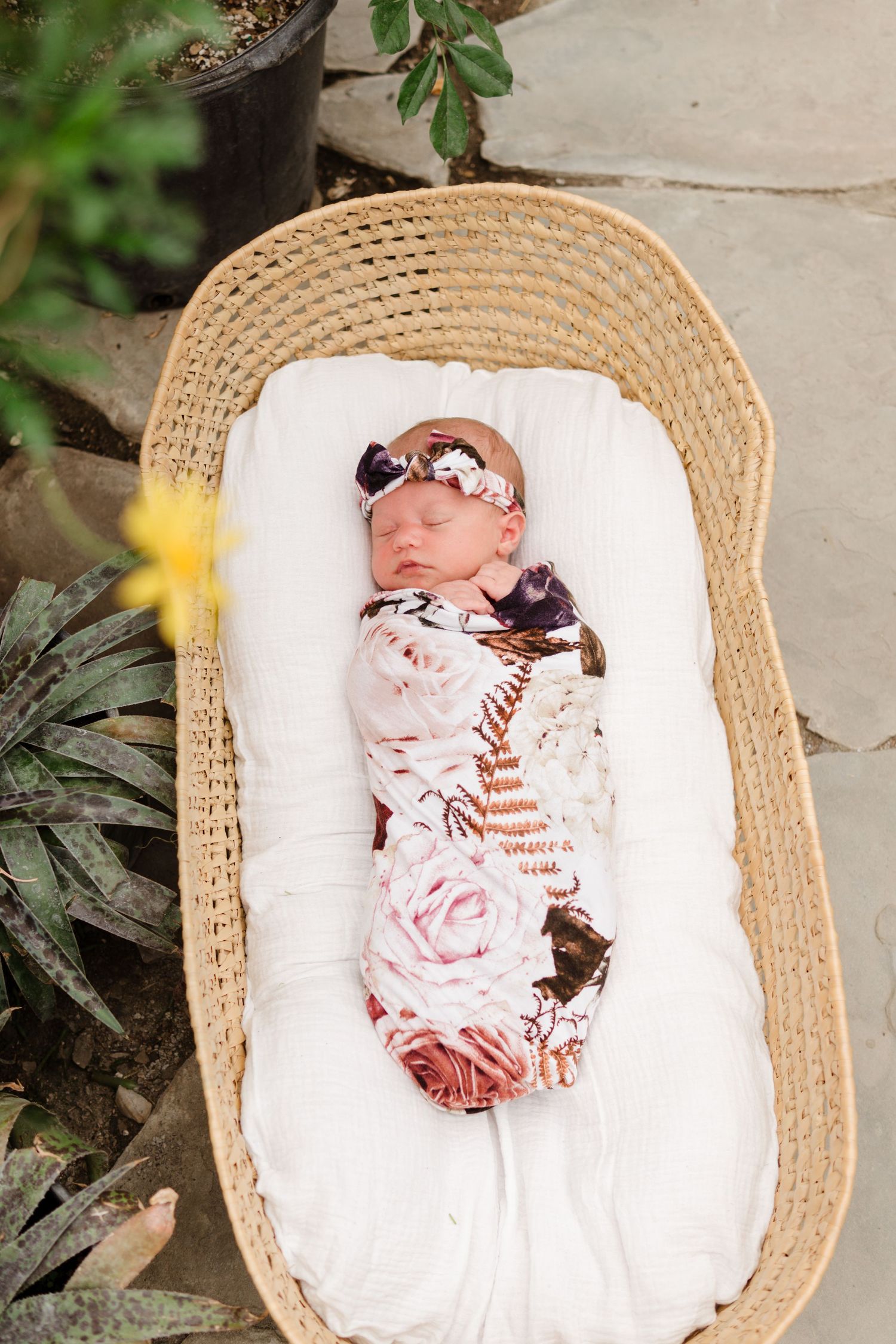 Newborn baby swaddled in floral wrap with matching headband, sleeping peacefully in a wicker Moses basket outdoors.