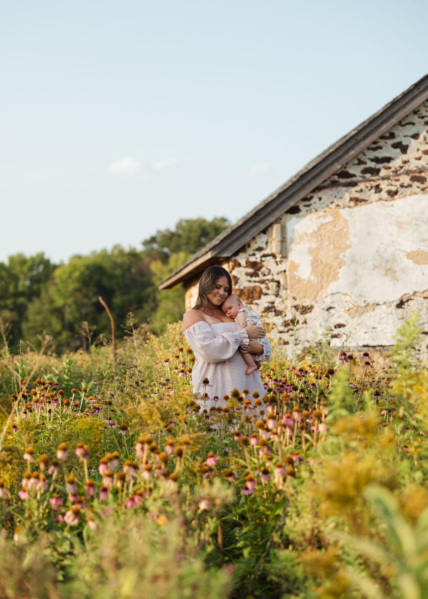 Woman in white off-shoulder dress holds baby in wildflower field beside rustic stone building on sunny day.