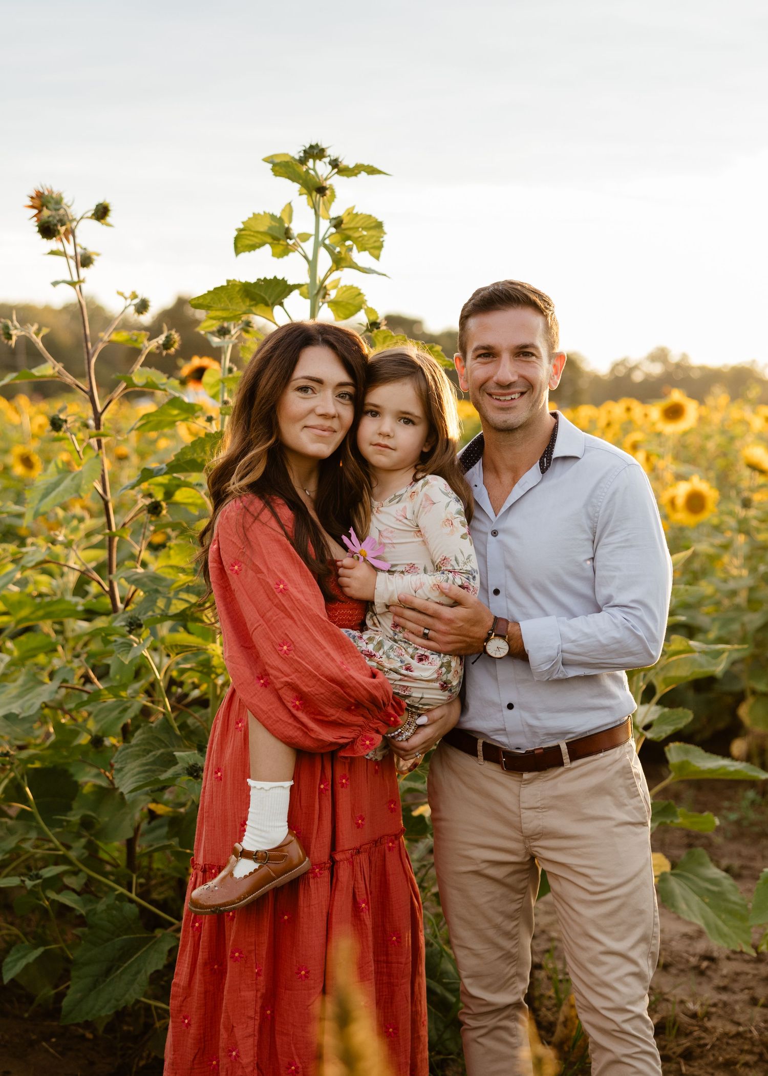 Family of three smiling in a sunflower field at golden hour, woman in red dress holding young child beside man in blue shirt.