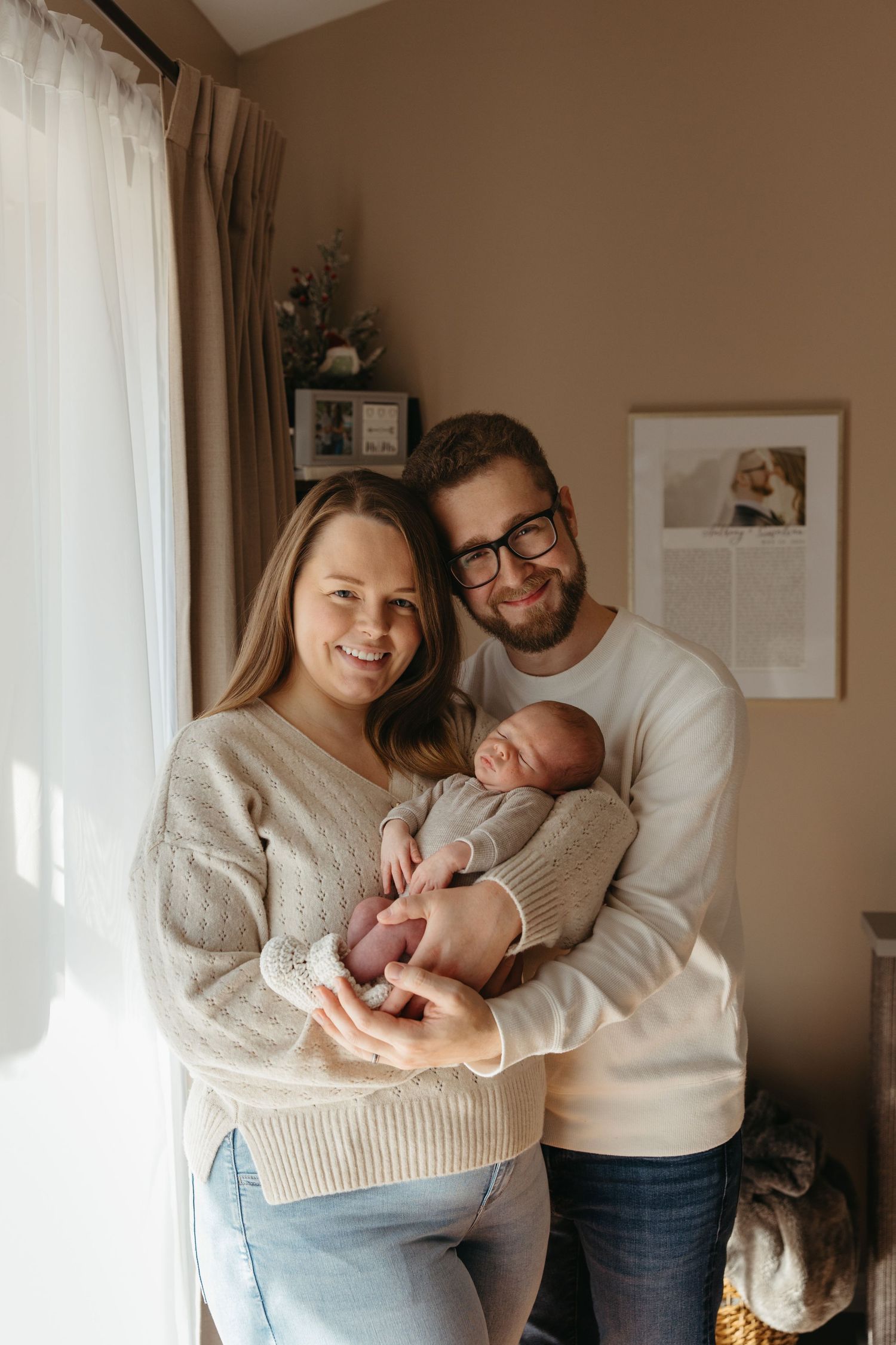 Smiling couple in cozy sweaters holding their newborn baby at home near a bright window.