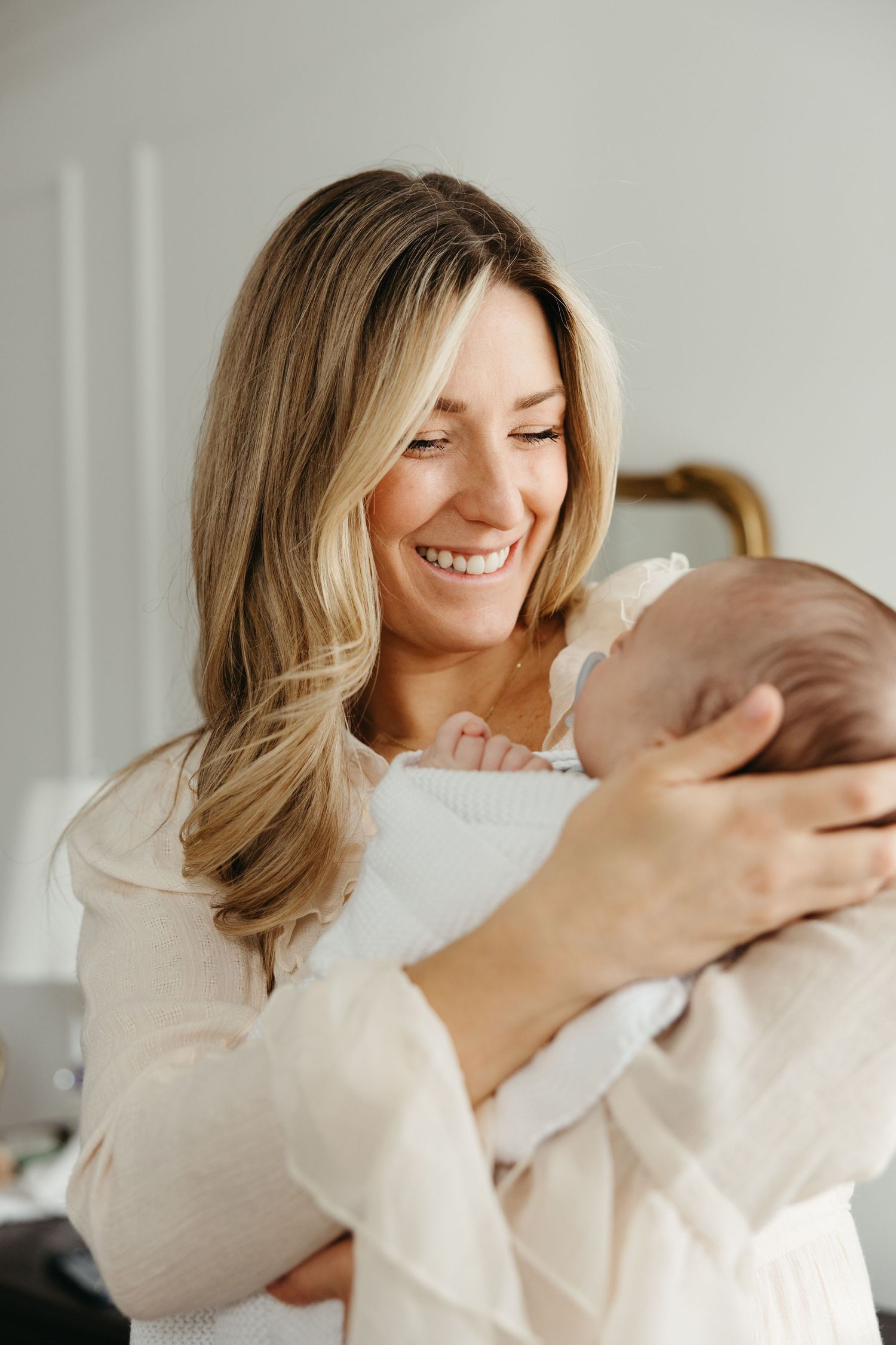 Joyful mother in cream sweater sharing intimate moments with newborn baby in minimalist white room setting.