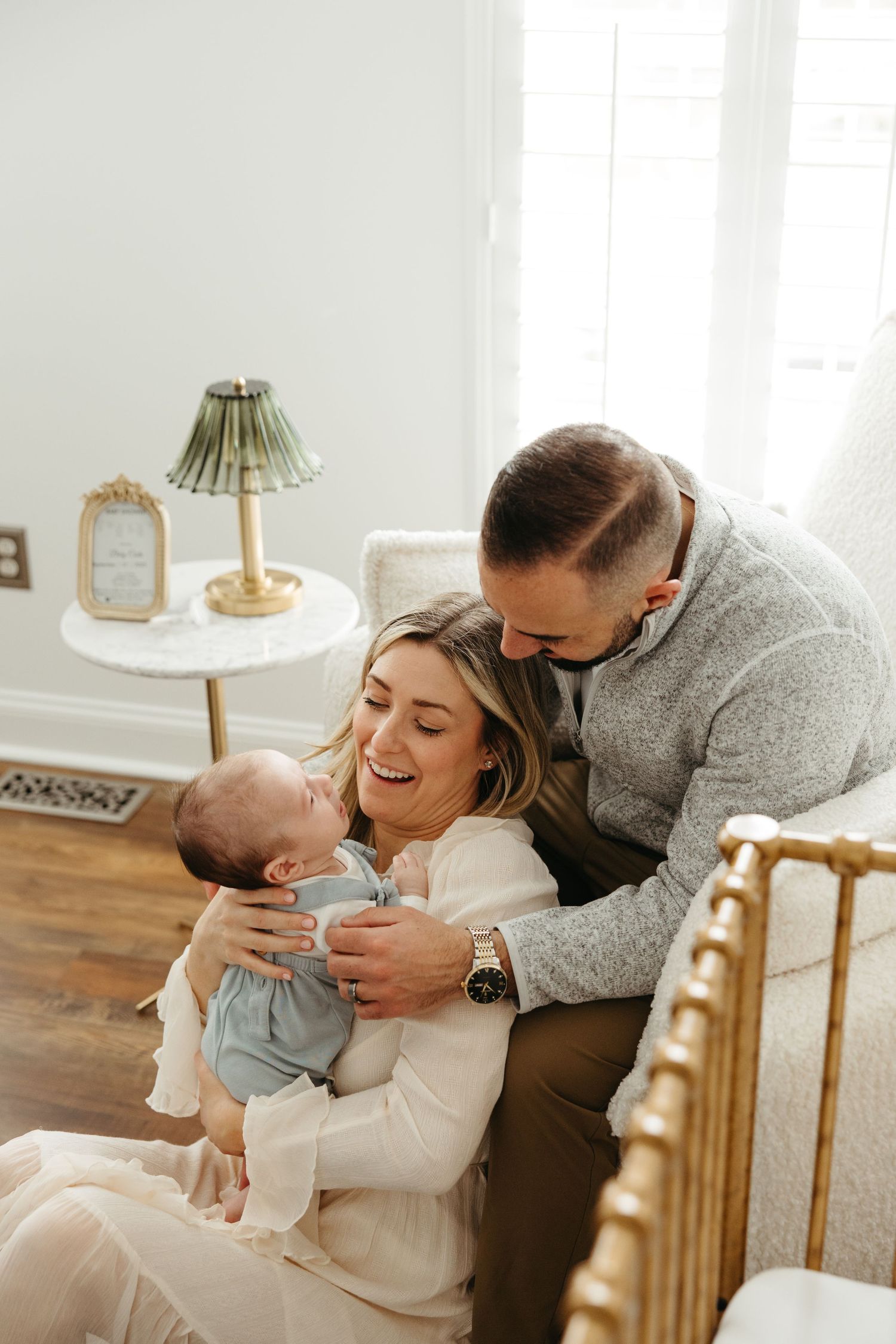 Tender family moment as parents in white and grey sweaters embrace their newborn baby in a bright sunlit room.