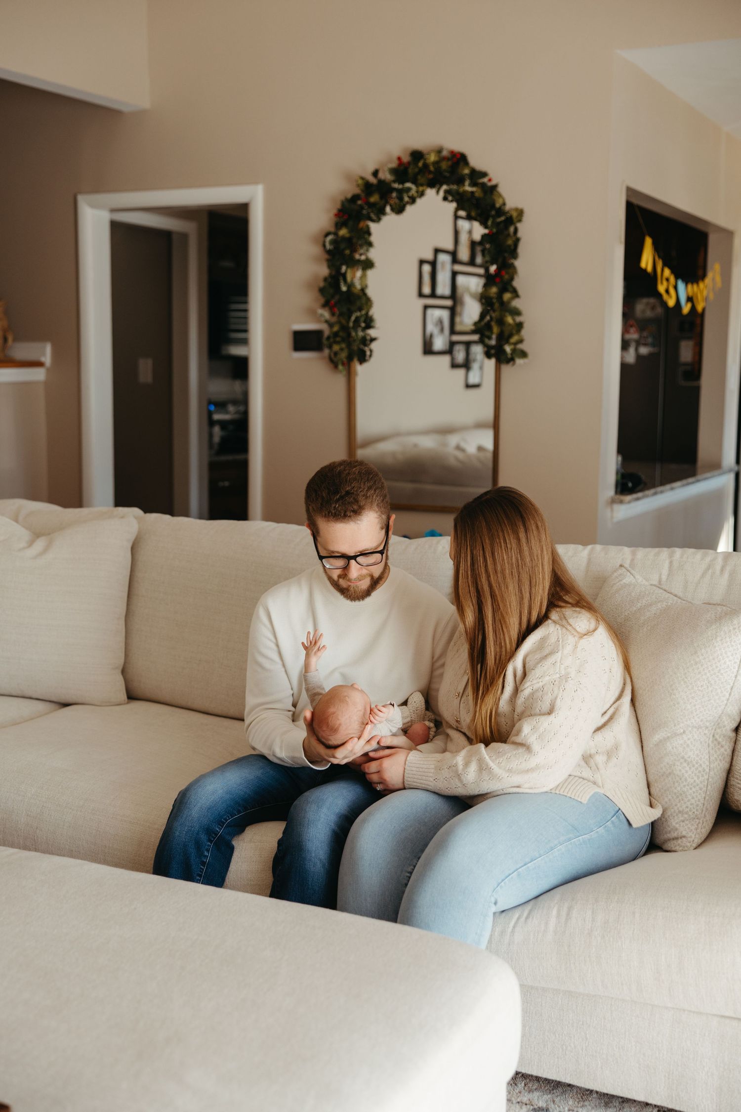 A young couple sits on a cream sofa, lovingly cradling their newborn baby in a cozy home living room.