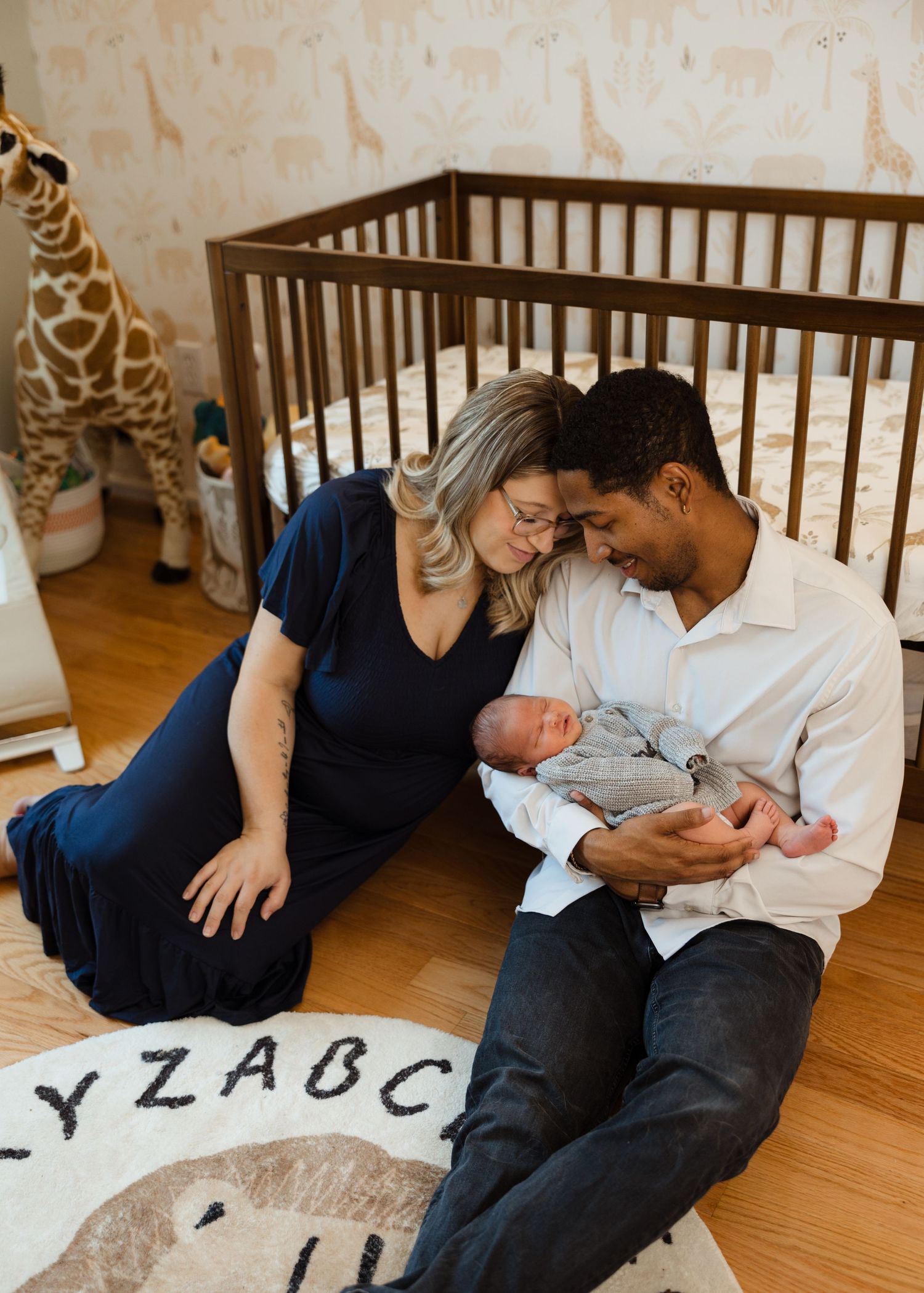 New parents sit on nursery floor with newborn baby, wooden crib and giraffe stuffed animal in background.