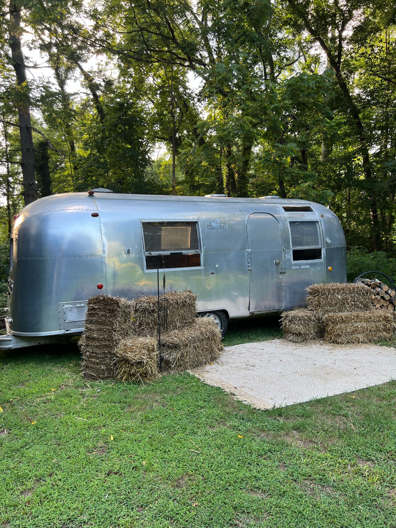 Vintage Airstream trailer parked outdoors surrounded by hay bales and lush green trees.