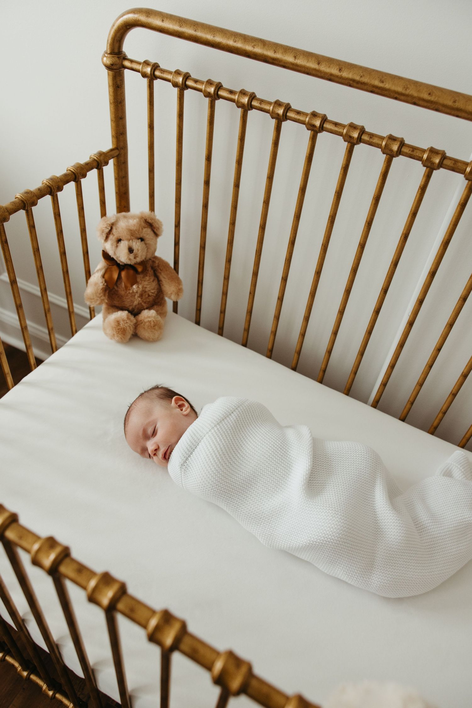 A small teddy bear sits atop white bedding in a wooden crib.