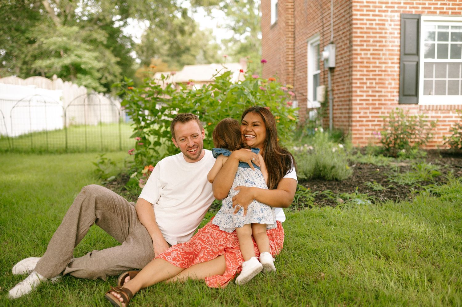 A group sits together on a grass lawn in front of a brick house on a sunny afternoon.