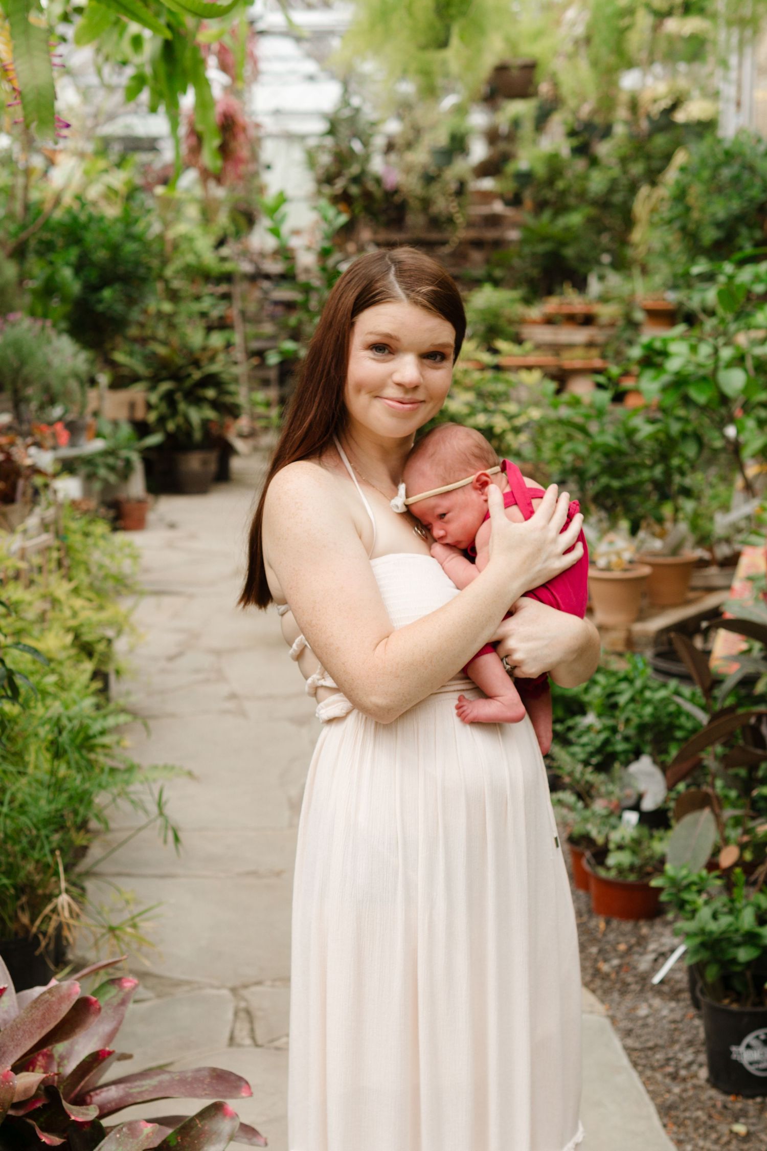 Young mother in white dress holds newborn baby in pink outfit in lush greenhouse garden setting.
