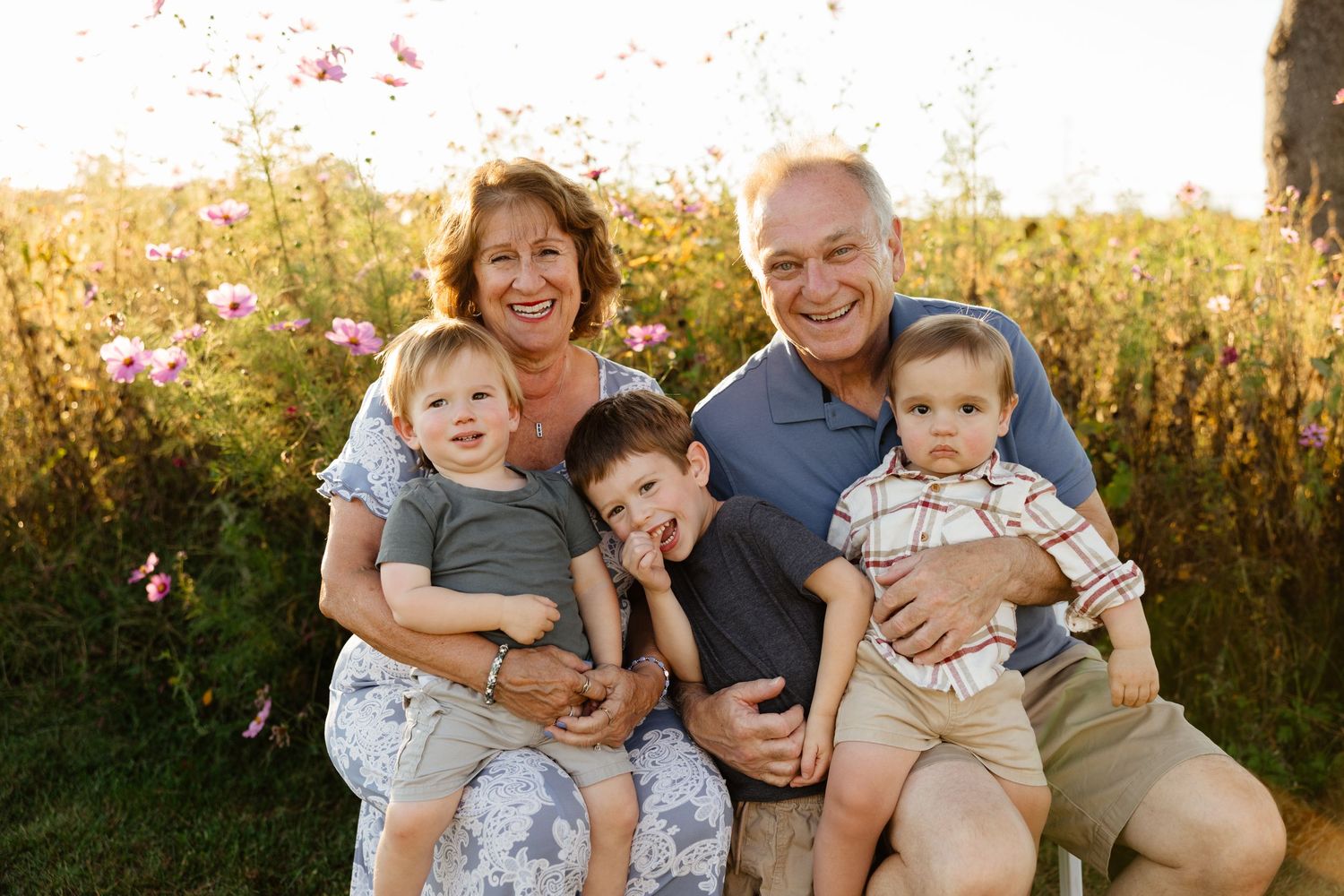 Grandparents smiling outdoors in a flower field, holding three young grandchildren in warm golden sunlight.