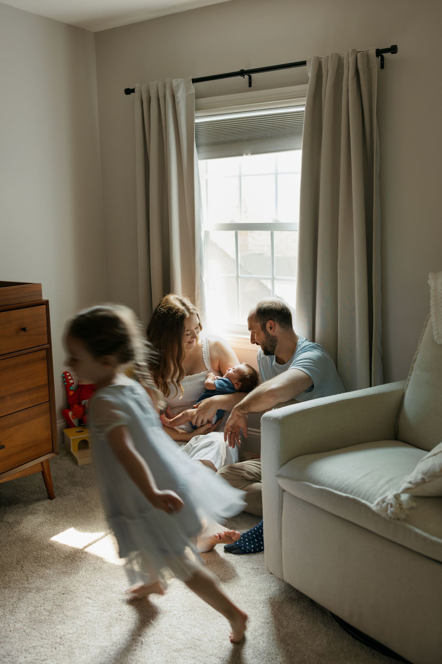 Parents sit by window with newborn while young girl runs playfully in sunlit nursery room.