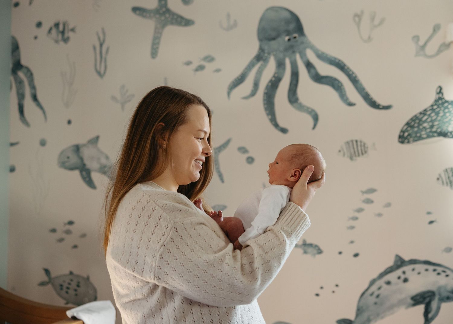 Mother smiling and holding newborn baby in nursery with ocean-themed wallpaper featuring octopus and sea creatures.