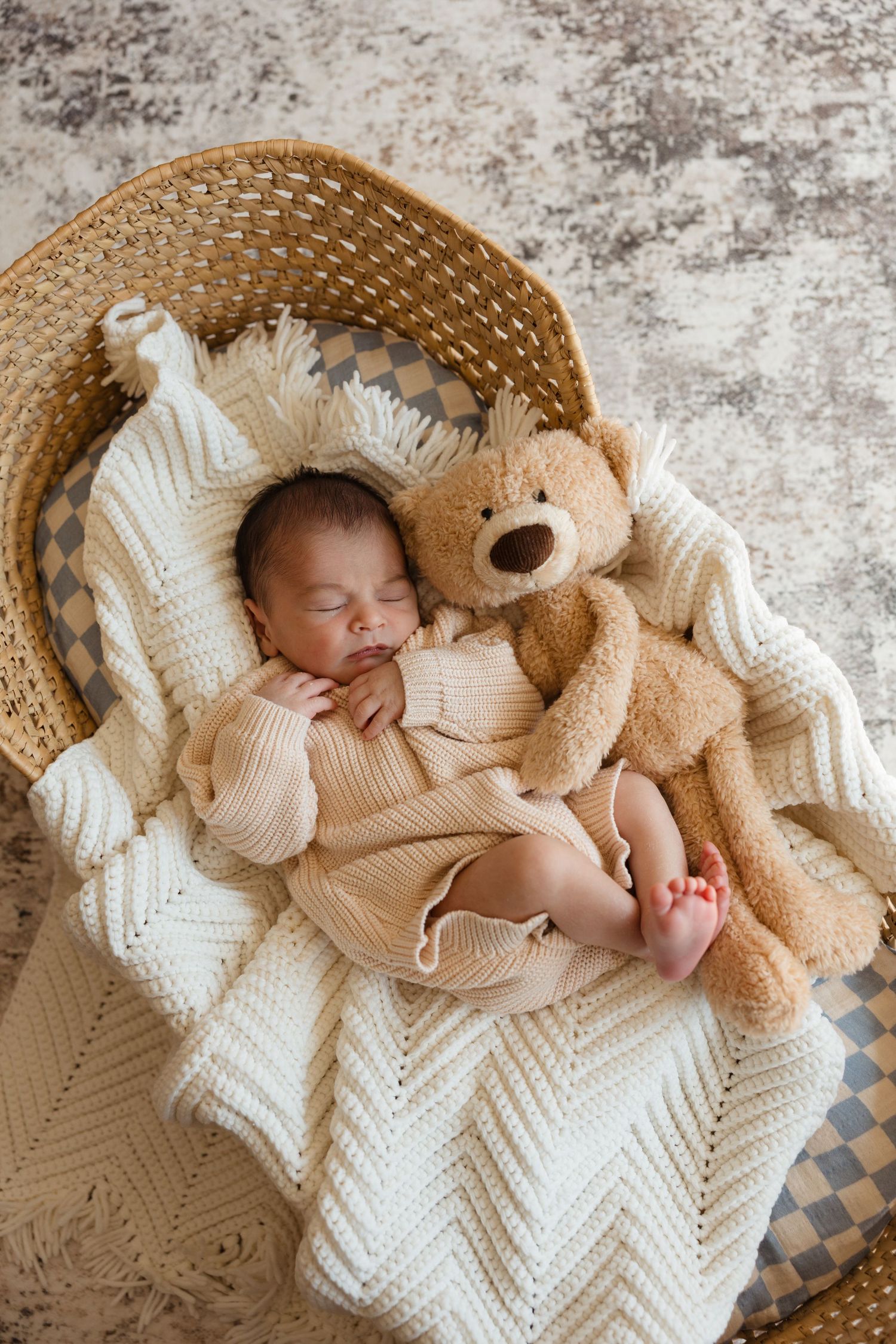 Sleeping newborn baby in beige knit outfit nestled in wicker basket with cream blanket and teddy bear.
