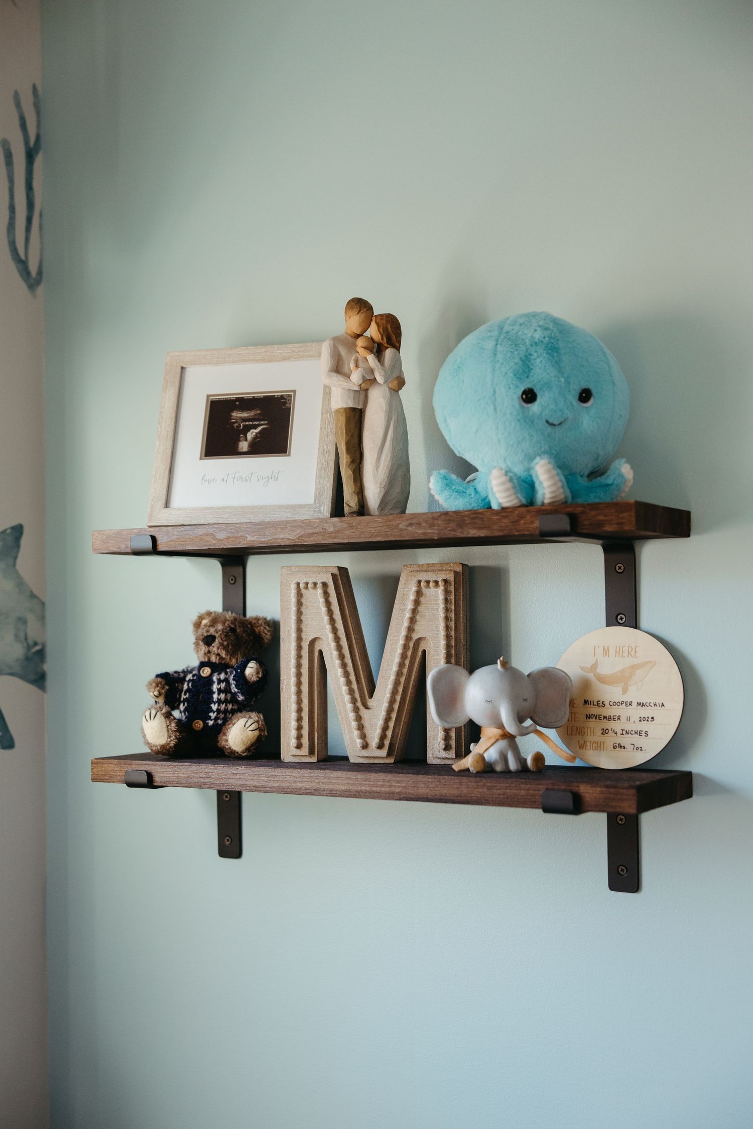 Two rustic wooden nursery shelves displaying a blue octopus plush, teddy bear, letter M, elephant toy, and framed ultrasound photo.