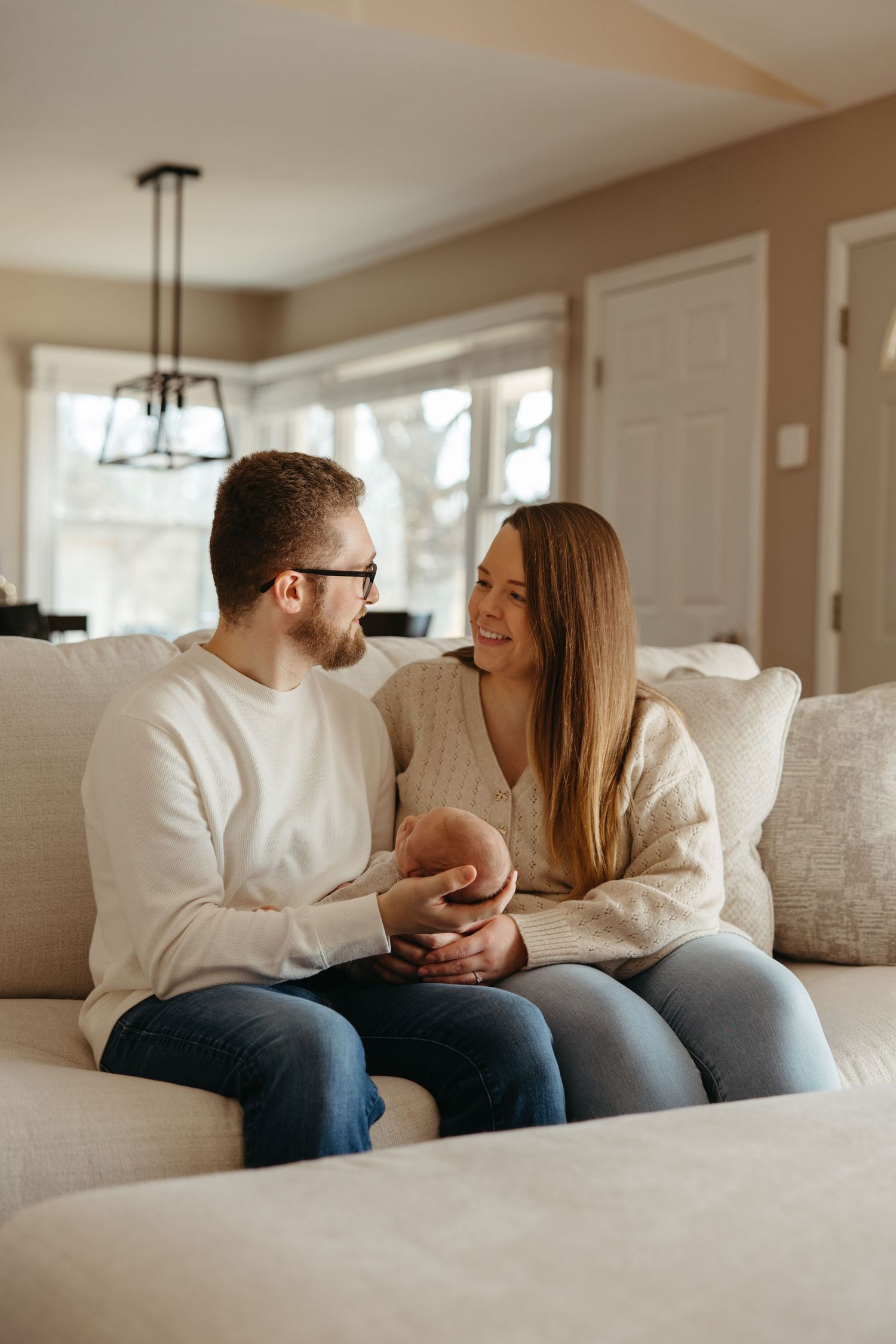 A couple sits together on a white sofa, smiling at each other and holding hands in a cozy living room.