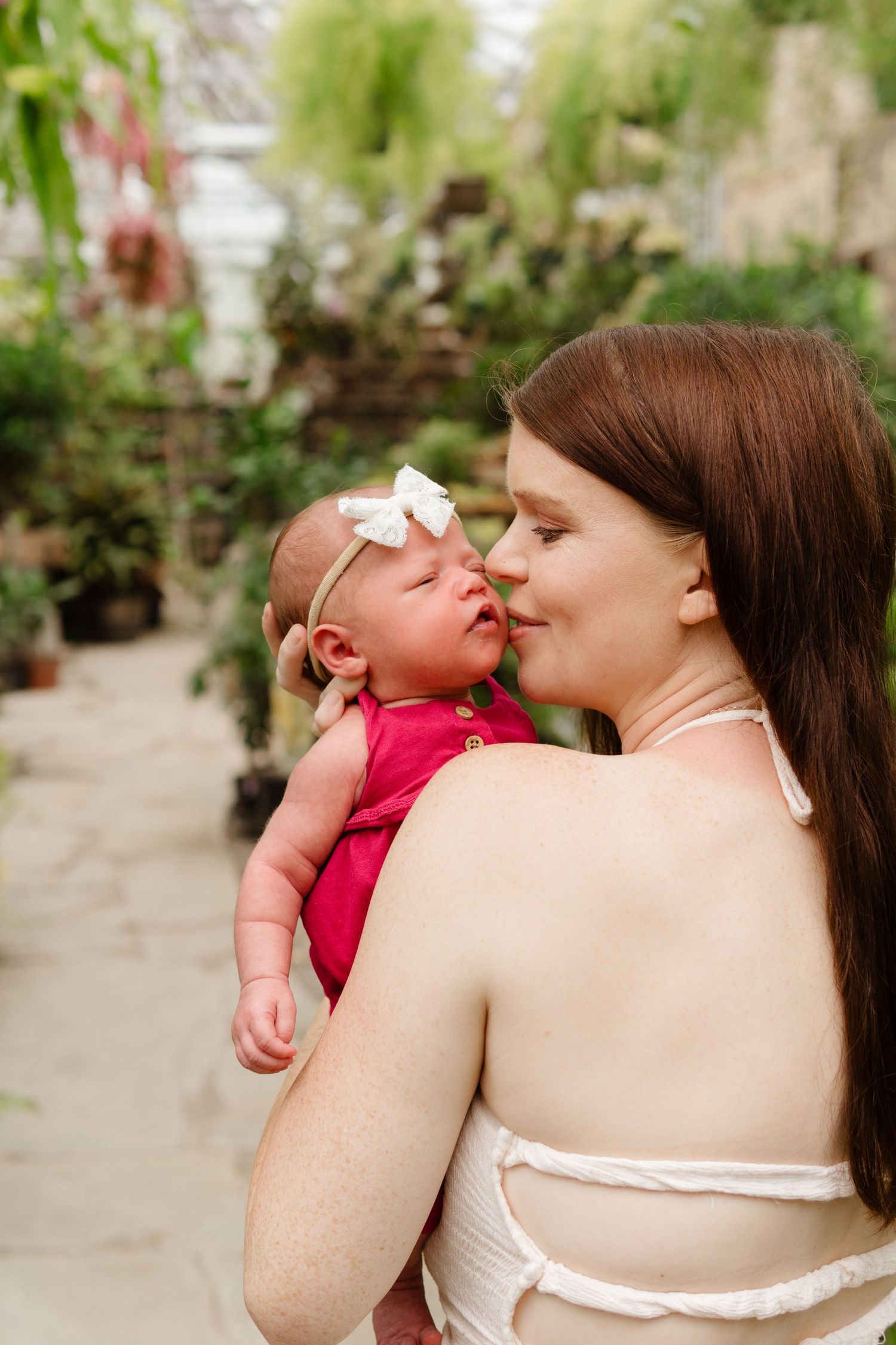 A mother tenderly kisses her newborn baby girl in a red outfit with a white bow headband in a lush greenhouse.