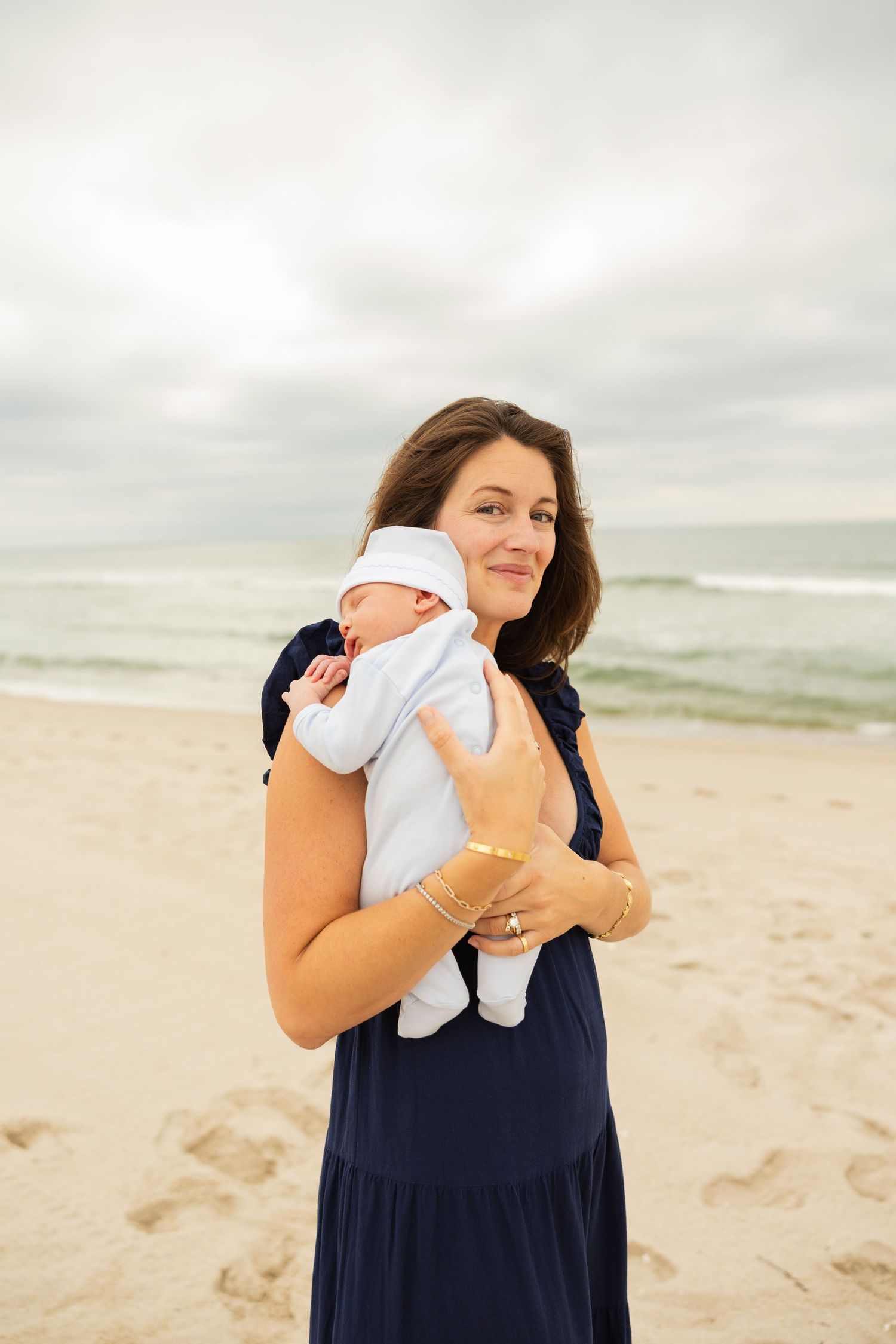 Woman in navy dress holds sleeping infant close while standing on sandy beach.