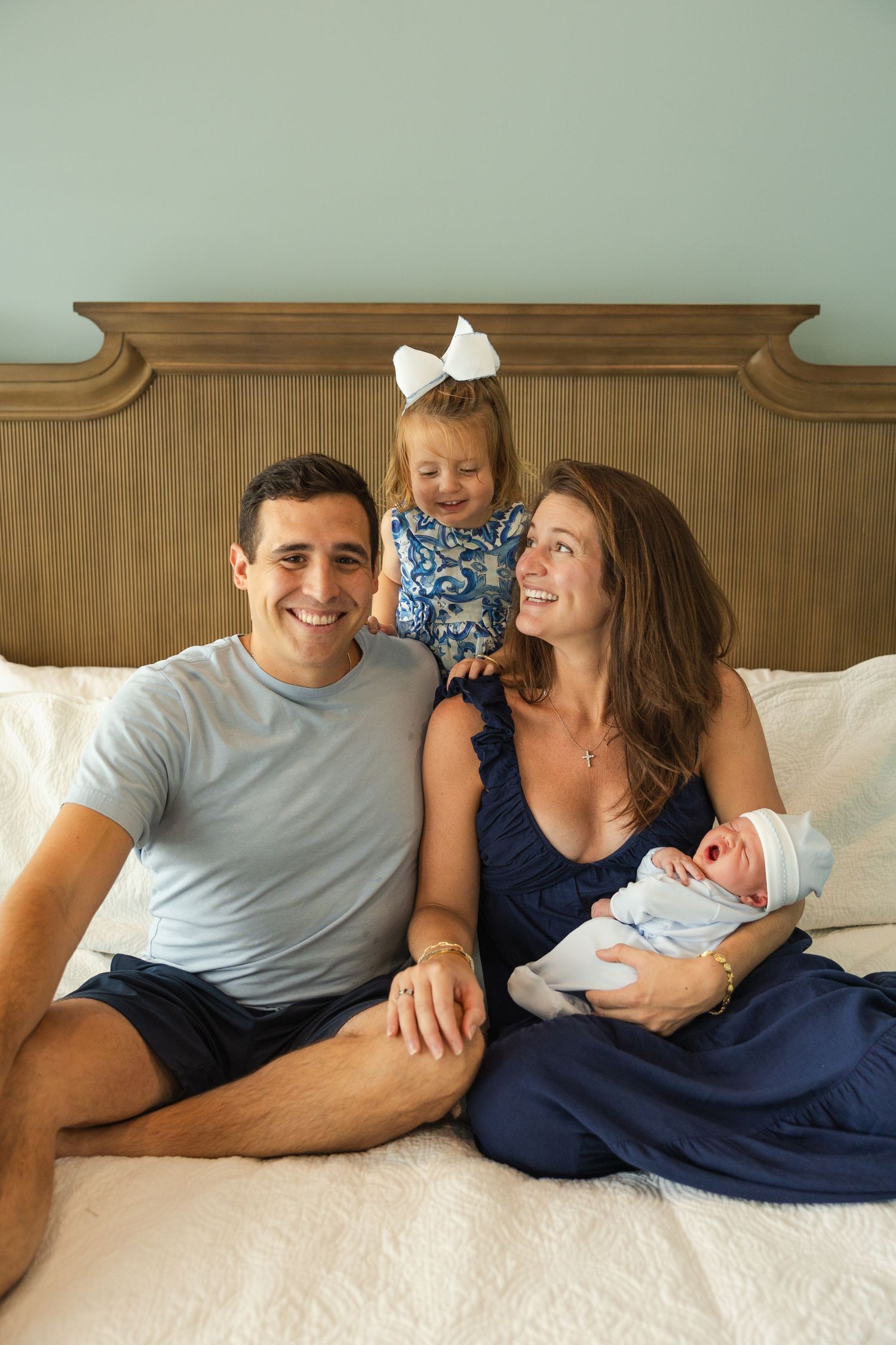 A family sits together on a bed with a wooden headboard, including a toddler in blue and a newborn baby.