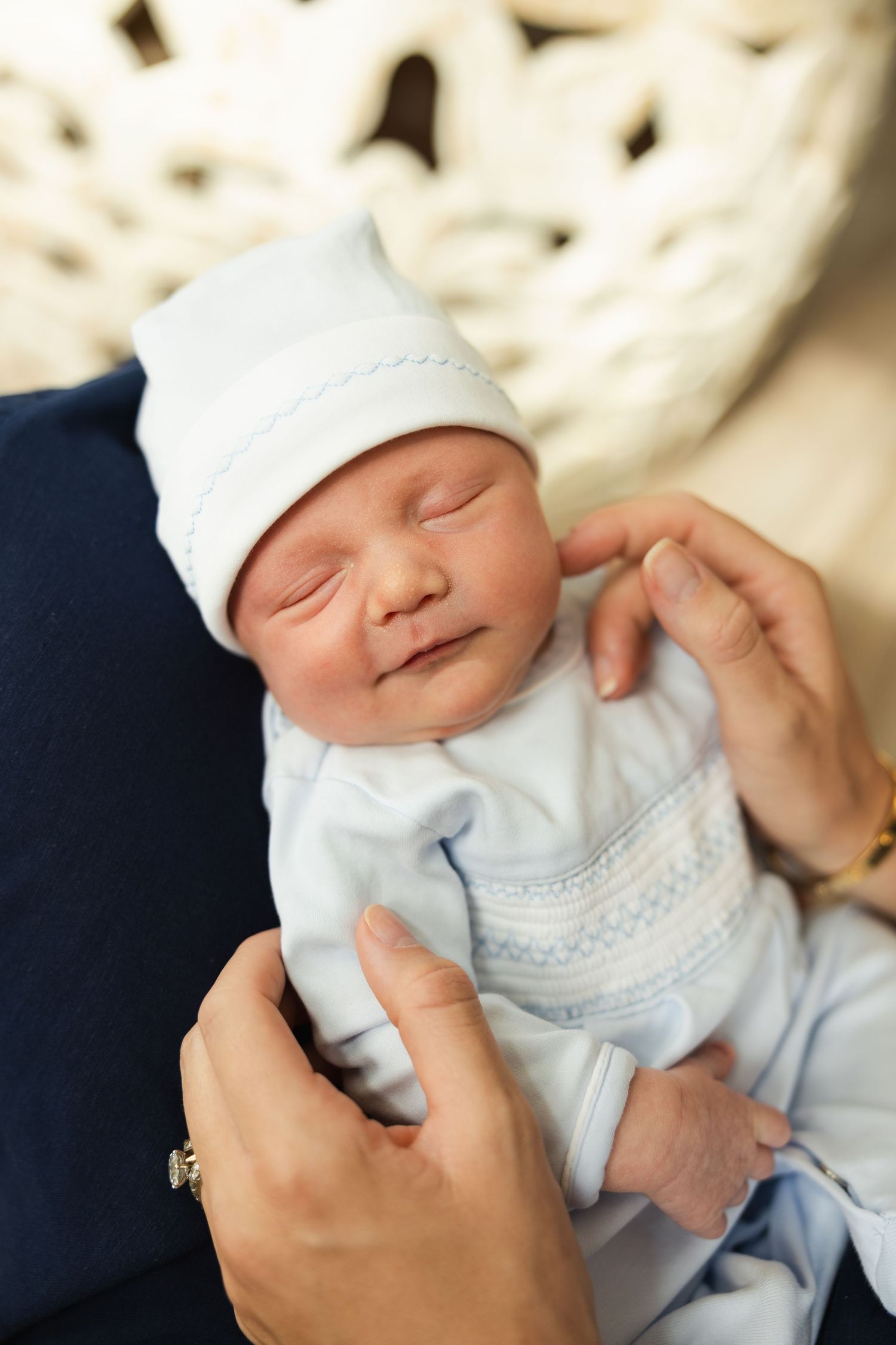 A sleeping newborn baby wearing a white cap and outfit smiles peacefully while being held.