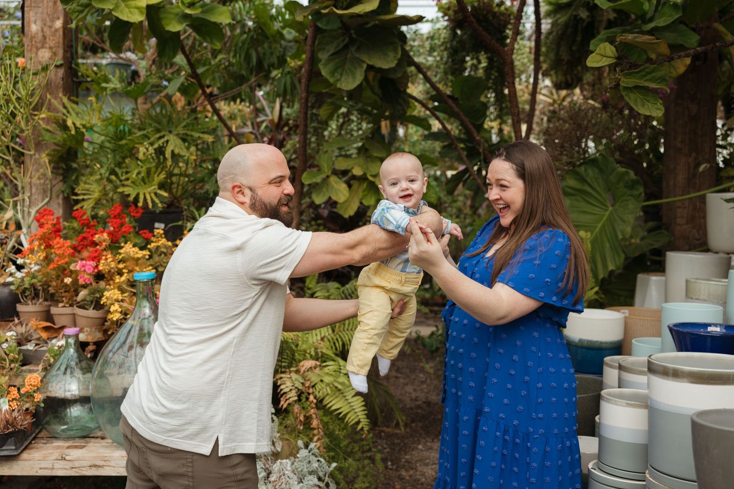 A happy family with a bald father and mother in blue dress lifting their smiling baby outdoors near lush greenery and pottery.