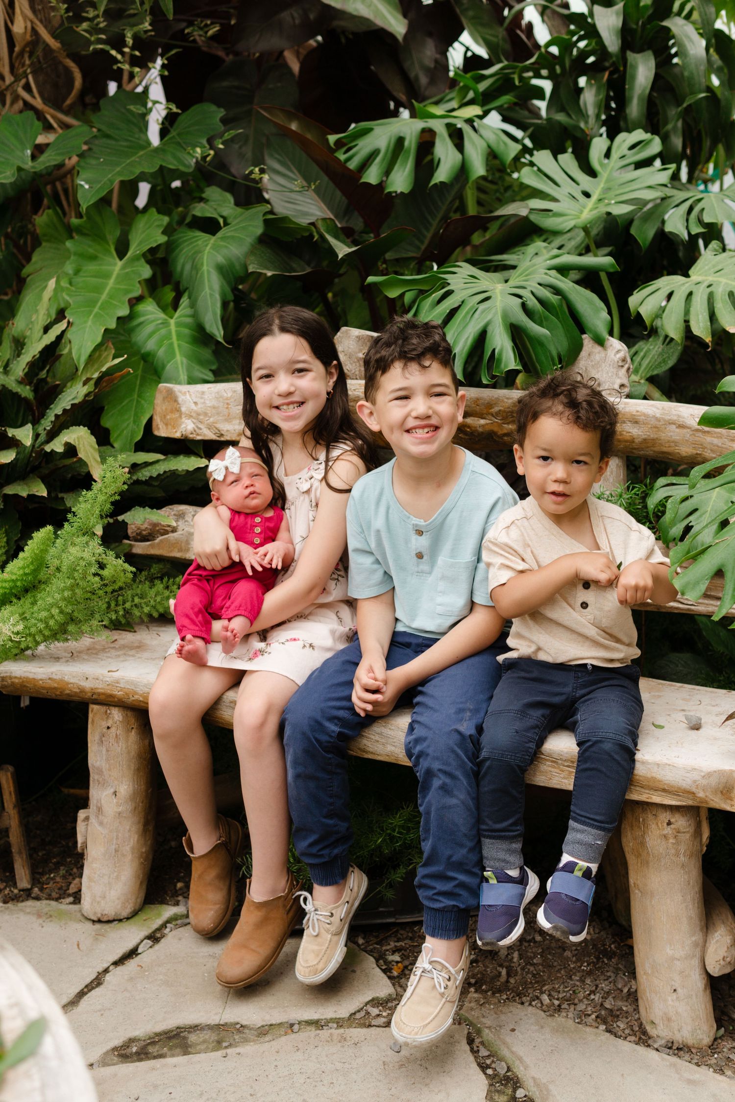 Three children sit smiling on a rustic wooden bench surrounded by tropical plants, with the oldest girl holding a baby doll.