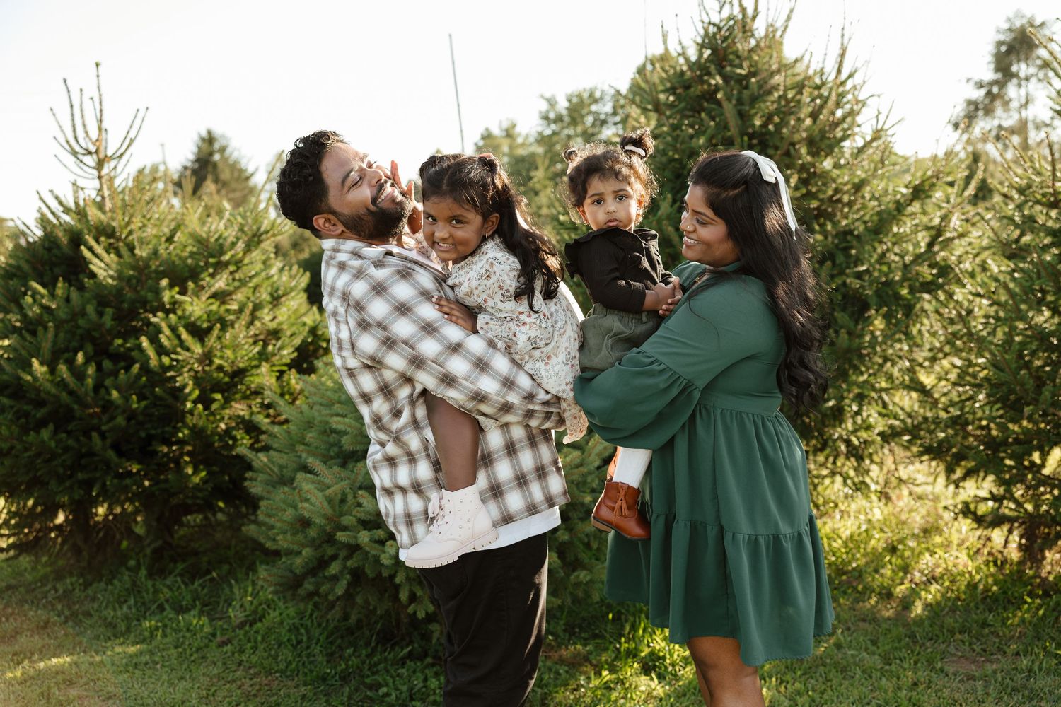 Family of four smiling at a Christmas tree farm, parents holding two young children among lush green trees.