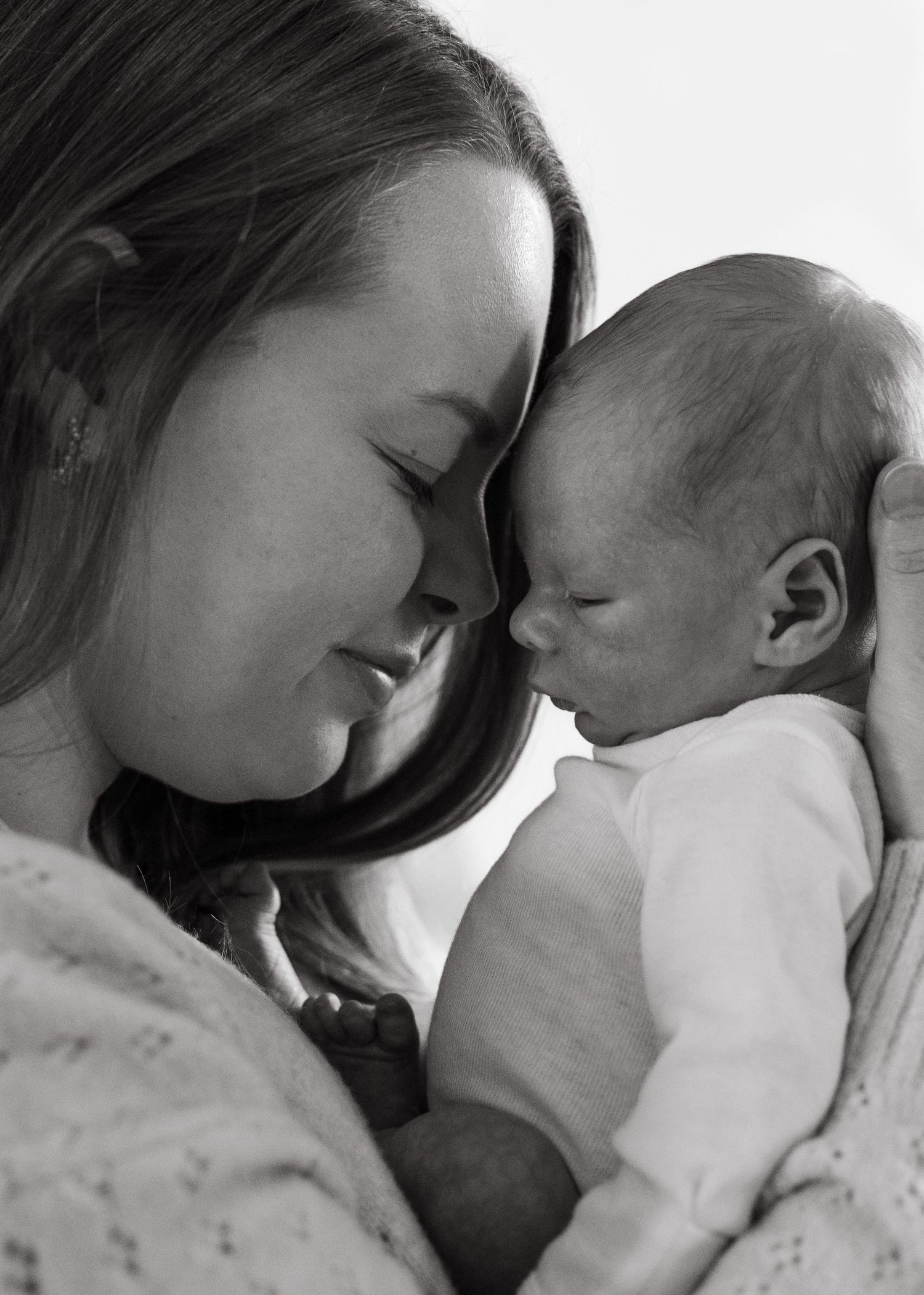 Black and white photo of a young mother tenderly holding her newborn baby, foreheads touching in an intimate moment.