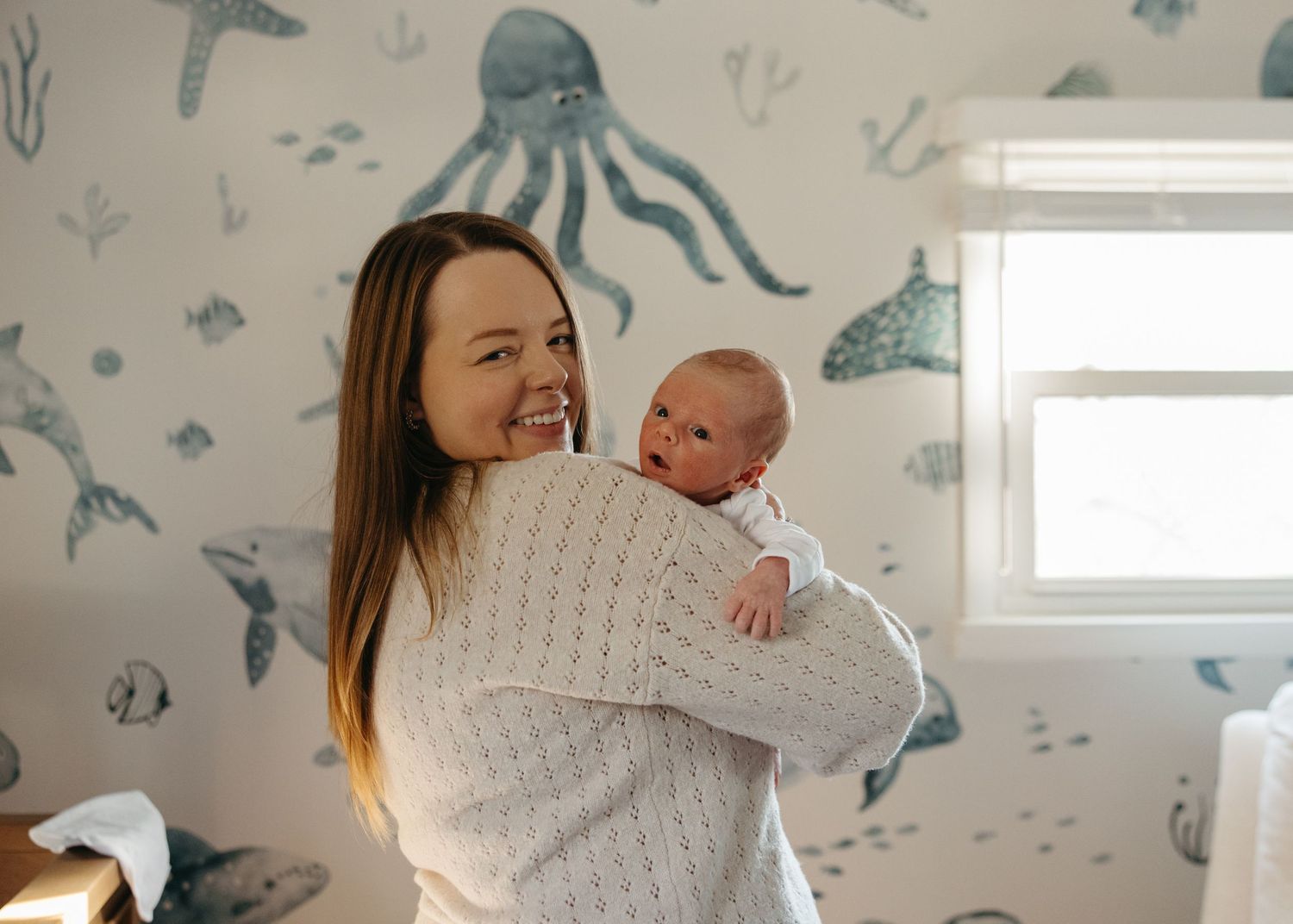 Smiling mother holding newborn baby in front of ocean-themed nursery wallpaper with octopus and fish.