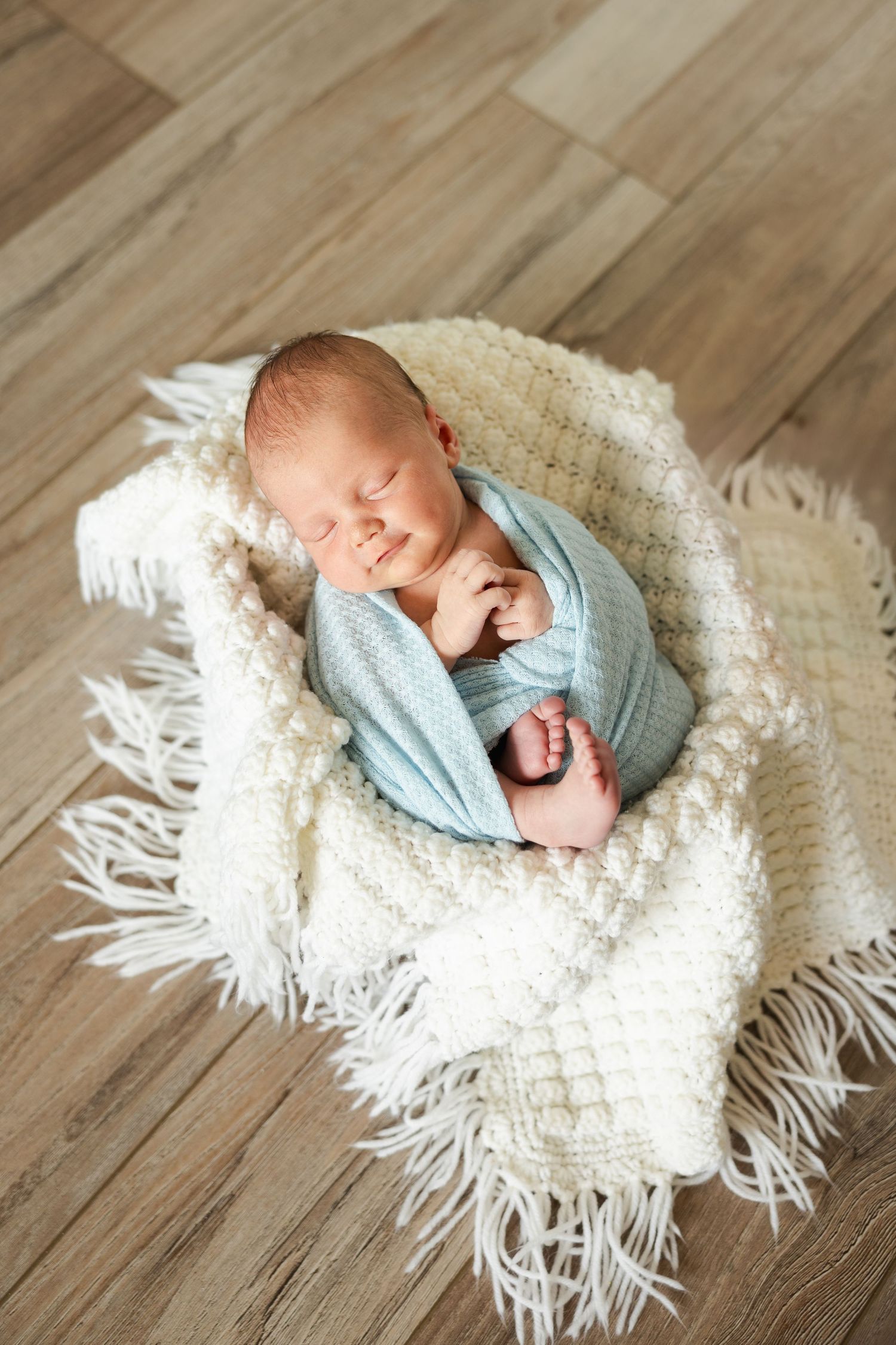 A sleeping newborn baby wrapped in a light blue sweater lies peacefully on a white knit blanket on wooden flooring.