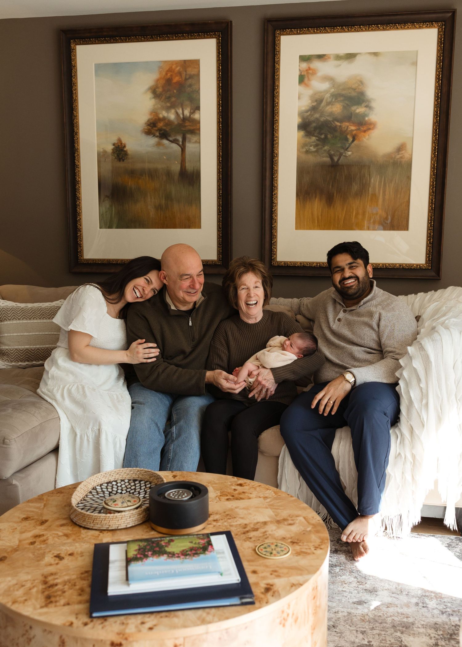 Multi-generational family smiling on couch with newborn baby, autumn landscape paintings on wall behind them.
