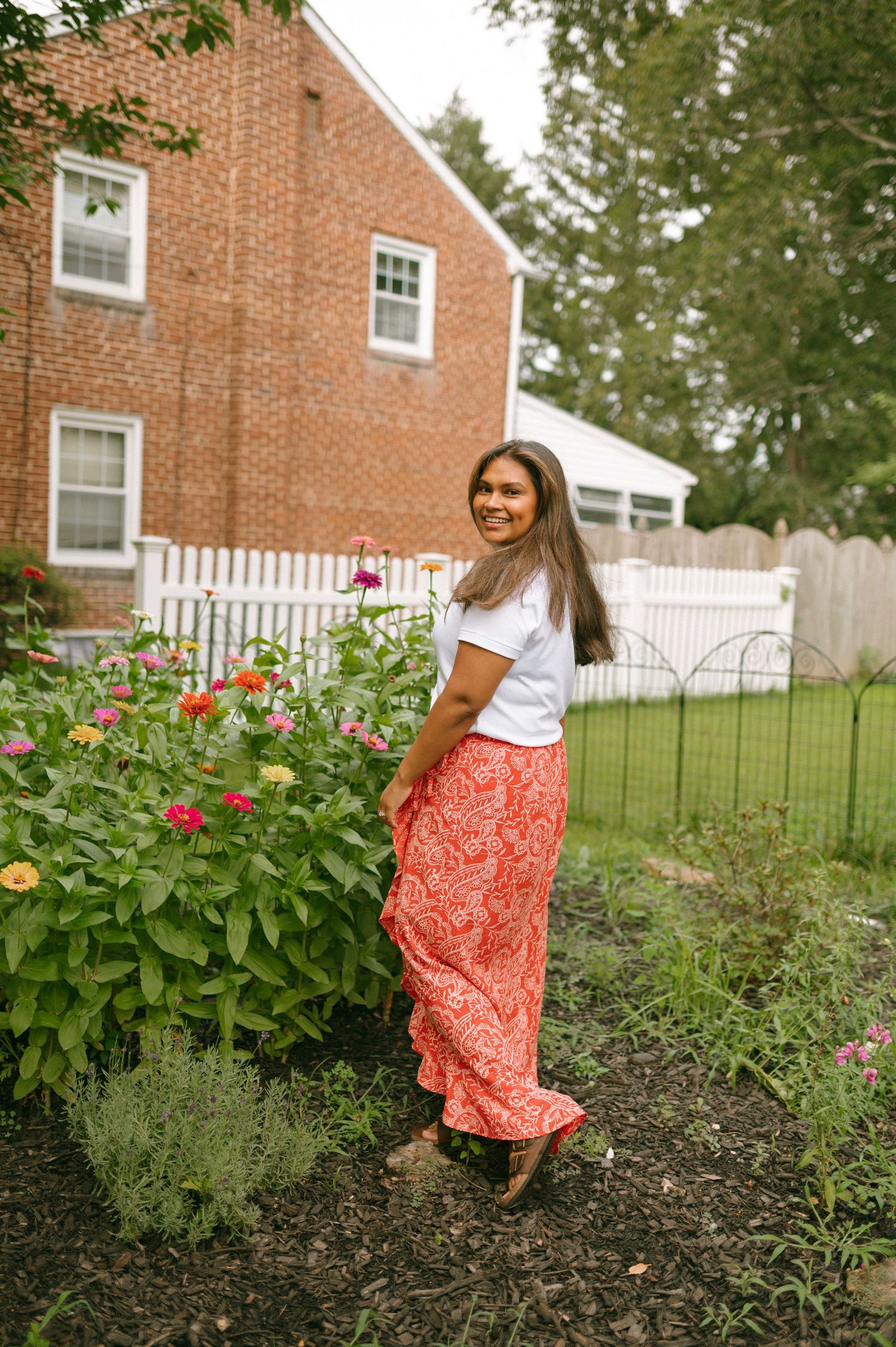 A person in a white top and coral floral skirt dances joyfully in a garden with blooming flowers and brick houses behind.