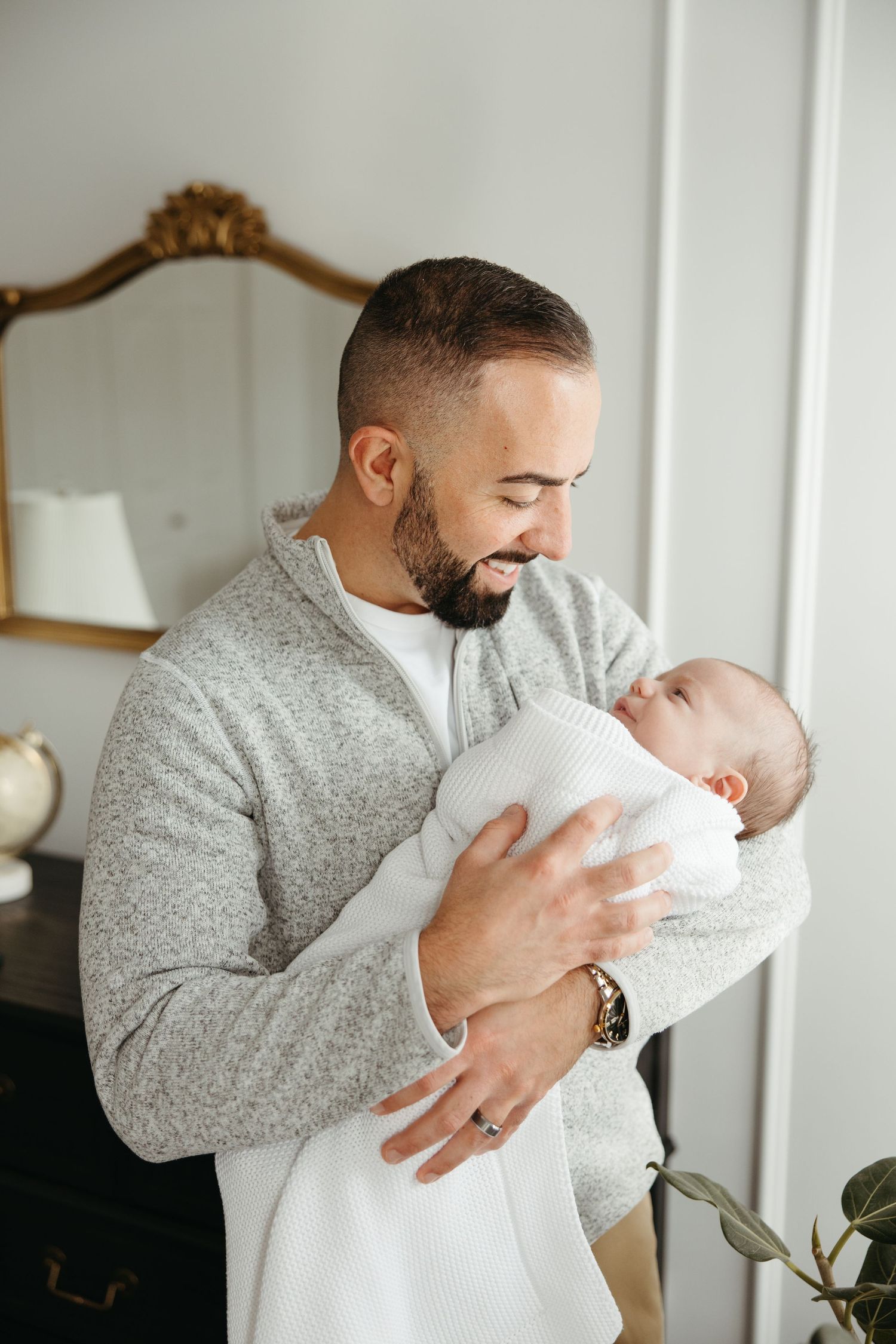 Father in grey sweater tenderly holds and kisses newborn baby wrapped in white blanket near window.