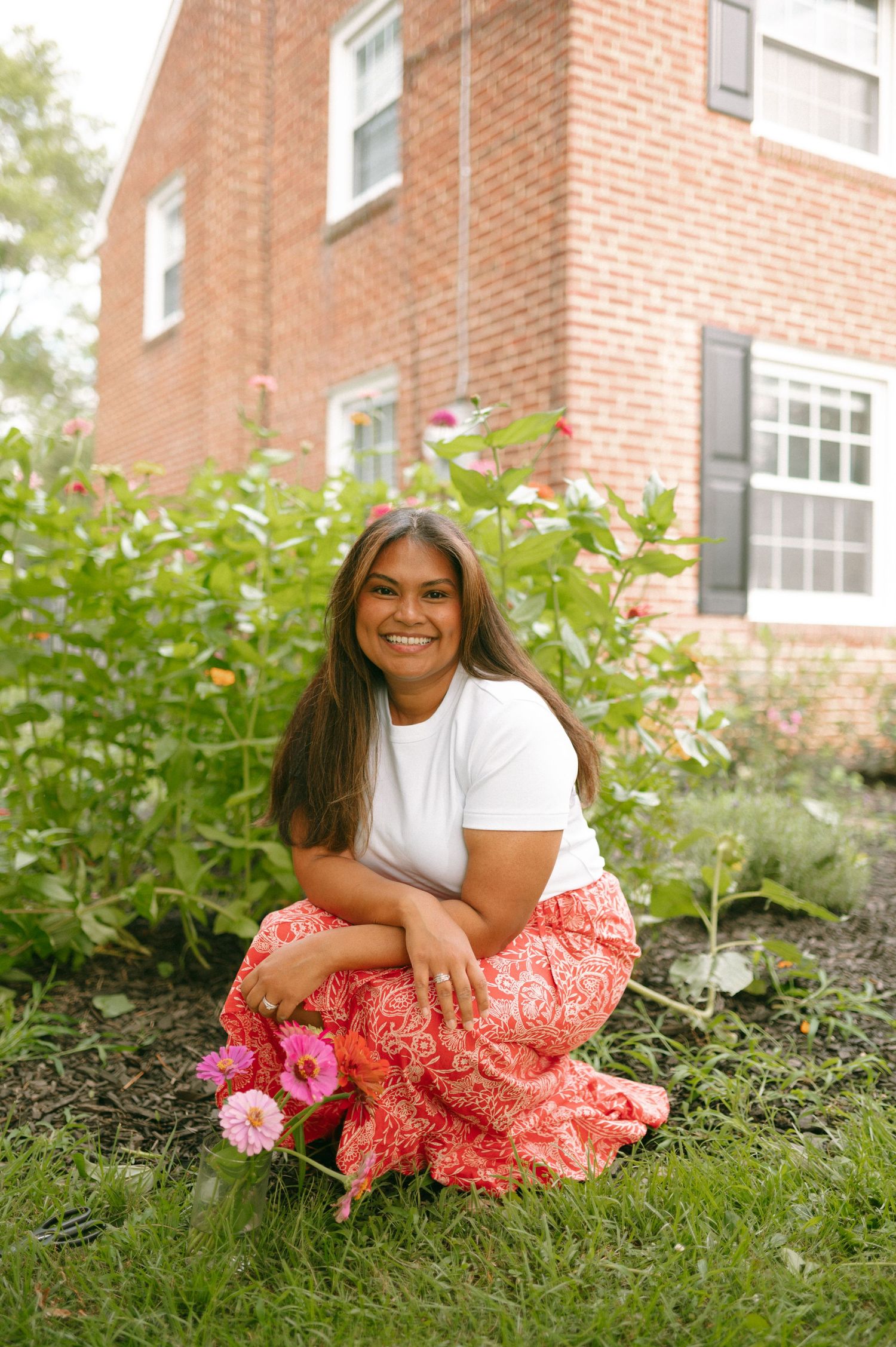 Someone in a white top and coral floral skirt poses casually on the grass in front of a brick building.