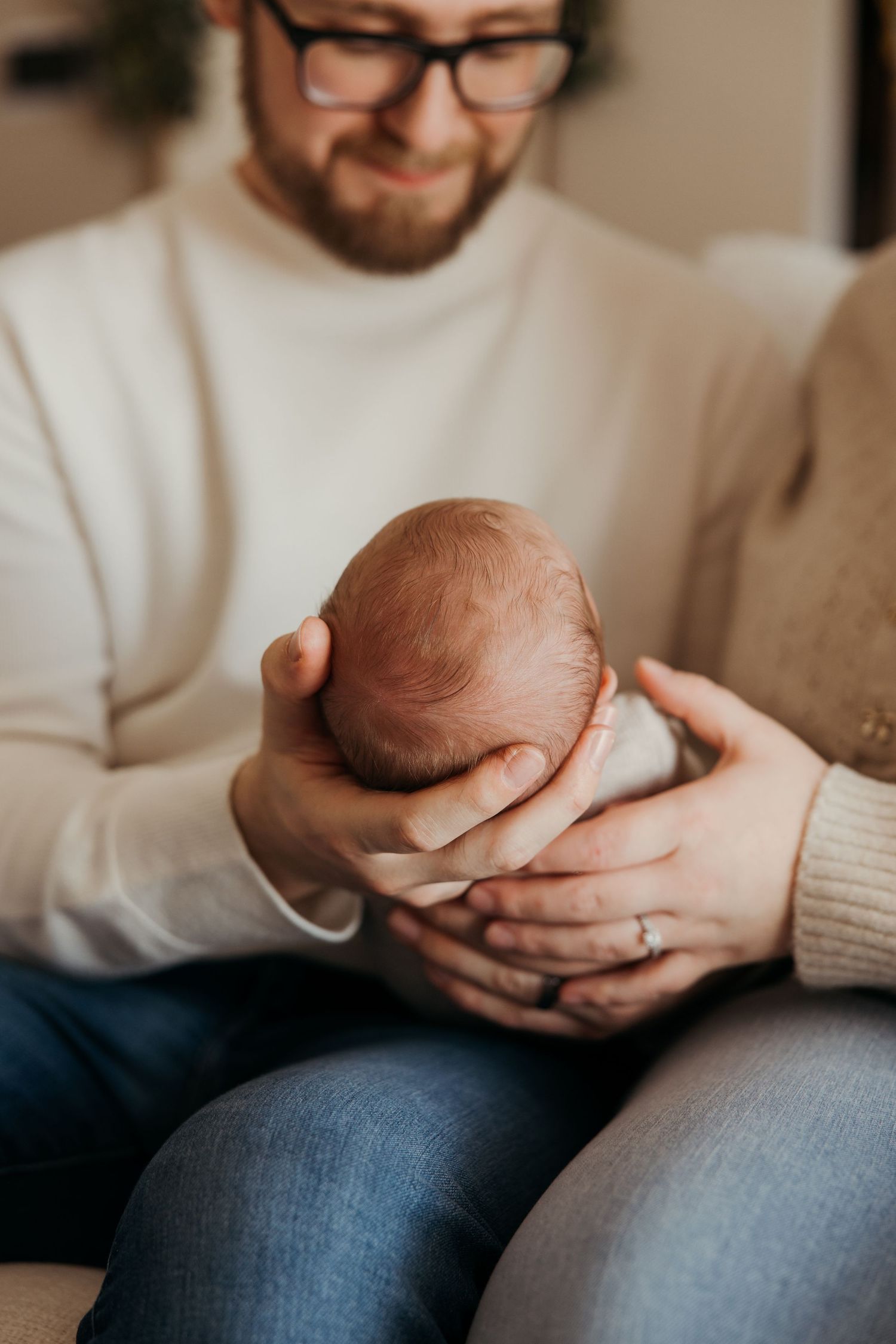 Parents tenderly cradling their newborn baby together, father in white sweater and jeans, mother wearing engagement ring.