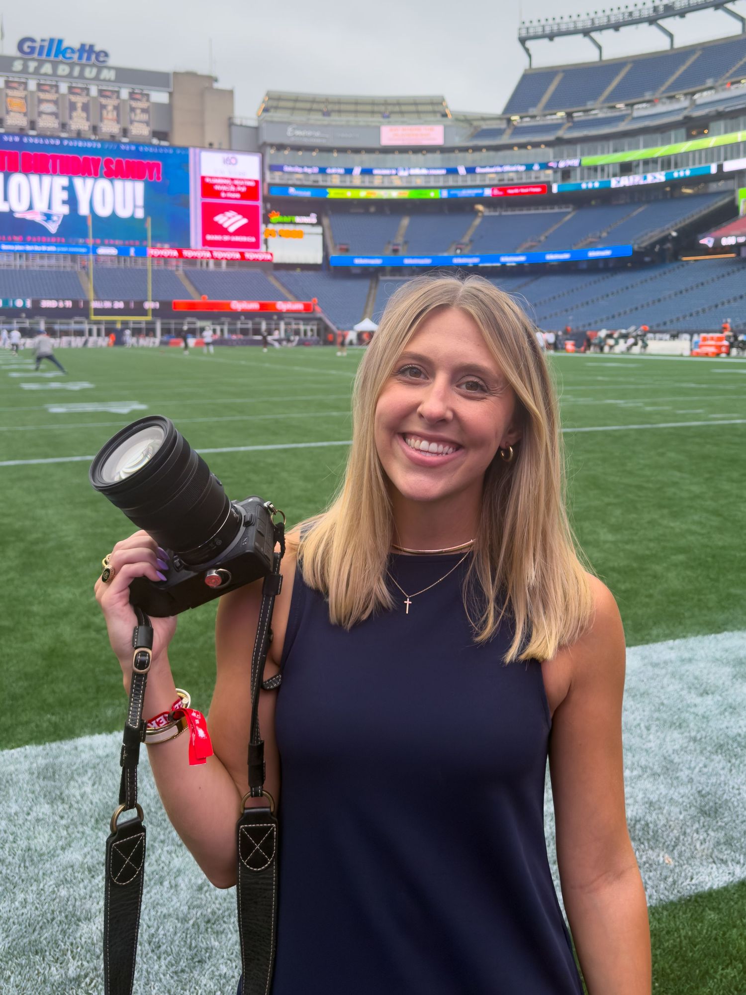 A smiling blonde woman holds a professional camera with telephoto lens on a football field at Gillette Stadium.