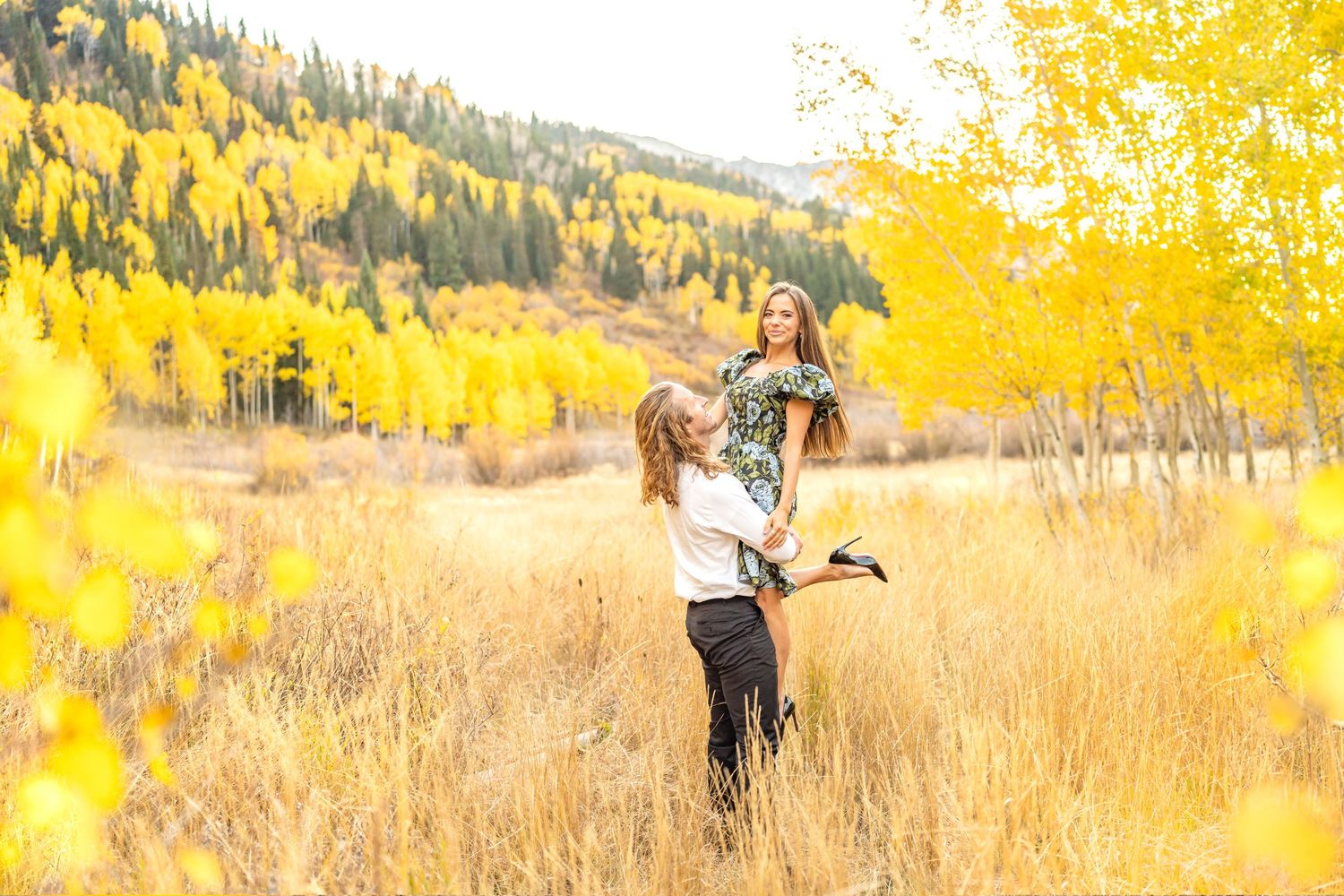 A couple embraces in a field surrounded by golden autumn foliage and tall grasses in the mountains.