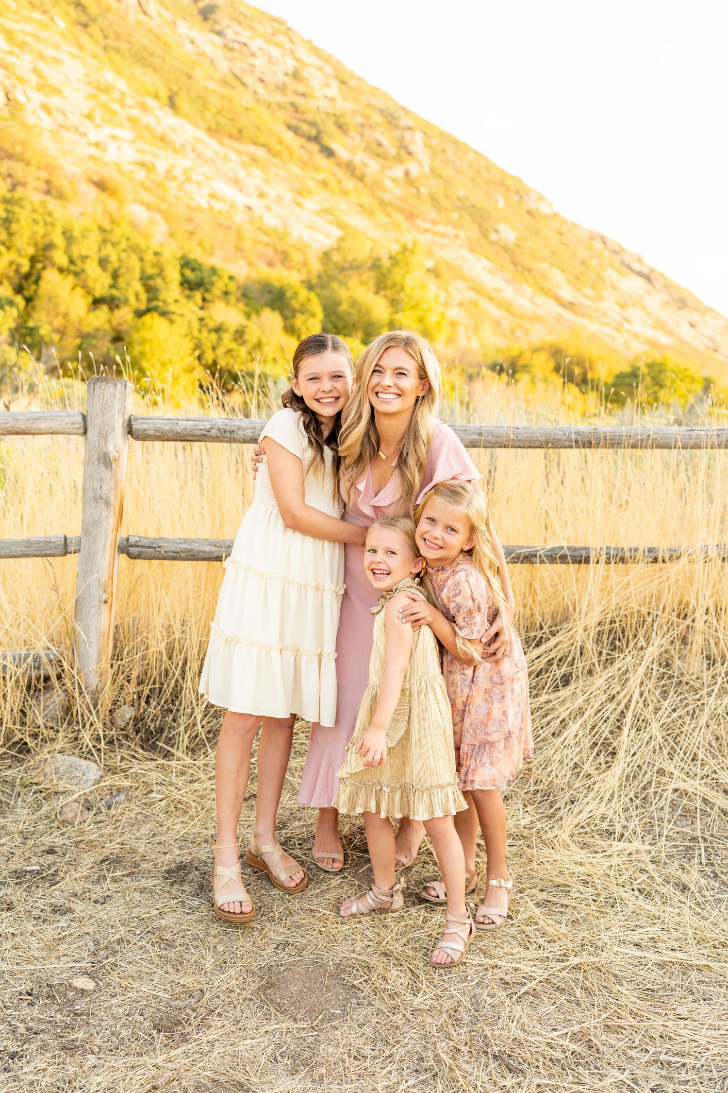 Three figures in light pink and beige dresses pose together against golden grass and mountain backdrop.