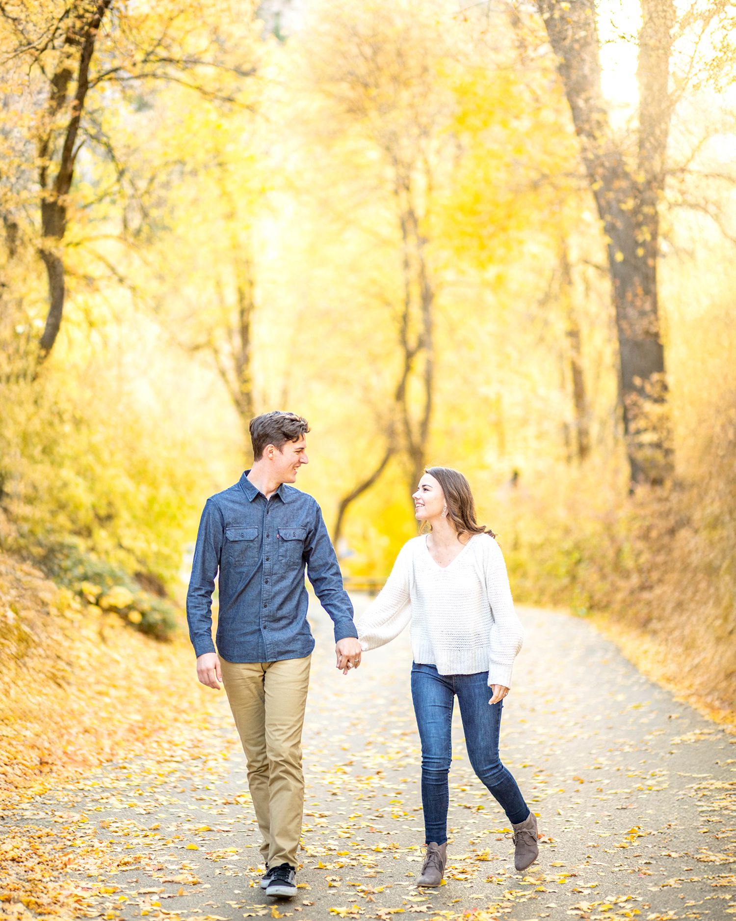 A couple walks hand in hand down a tree-lined path covered in golden fall foliage during autumn.