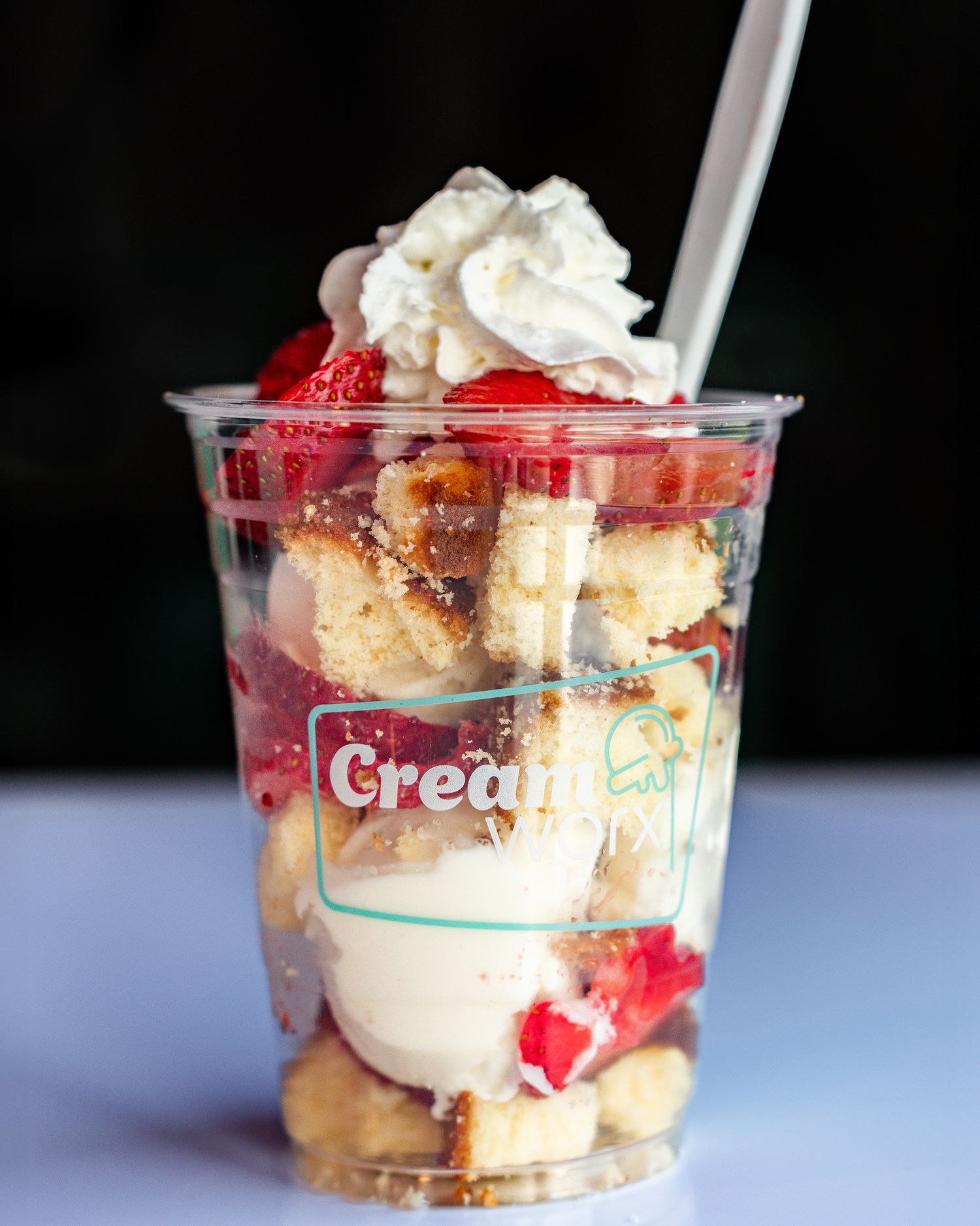 A layered strawberry poundcake ice cream sundae served in a clear plastic cup with whipped cream and strawberries.