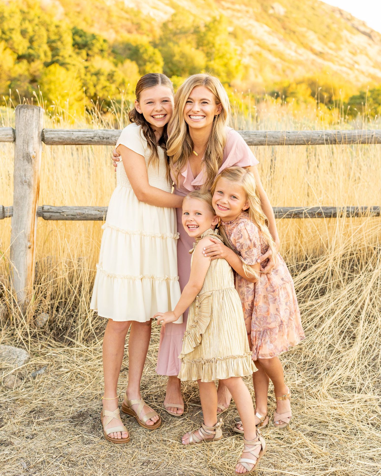 A family group dressed in soft pink and cream dresses poses together by a rustic wooden fence in golden evening light.