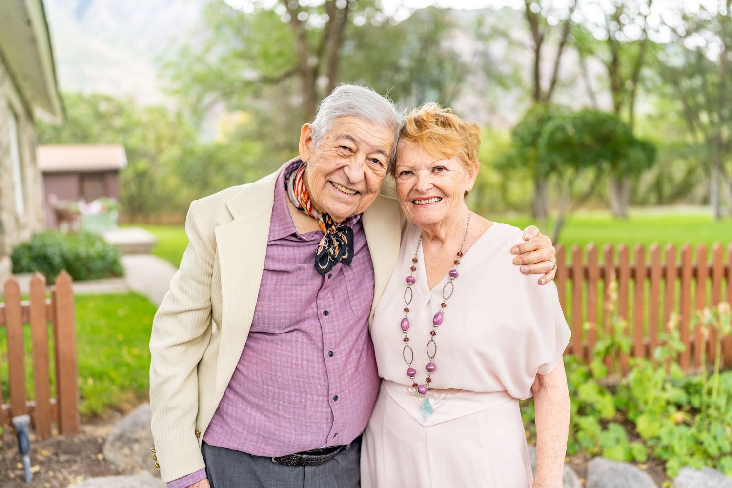 Two elderly people embrace and smile warmly while standing together in a garden with a wooden fence in the background.