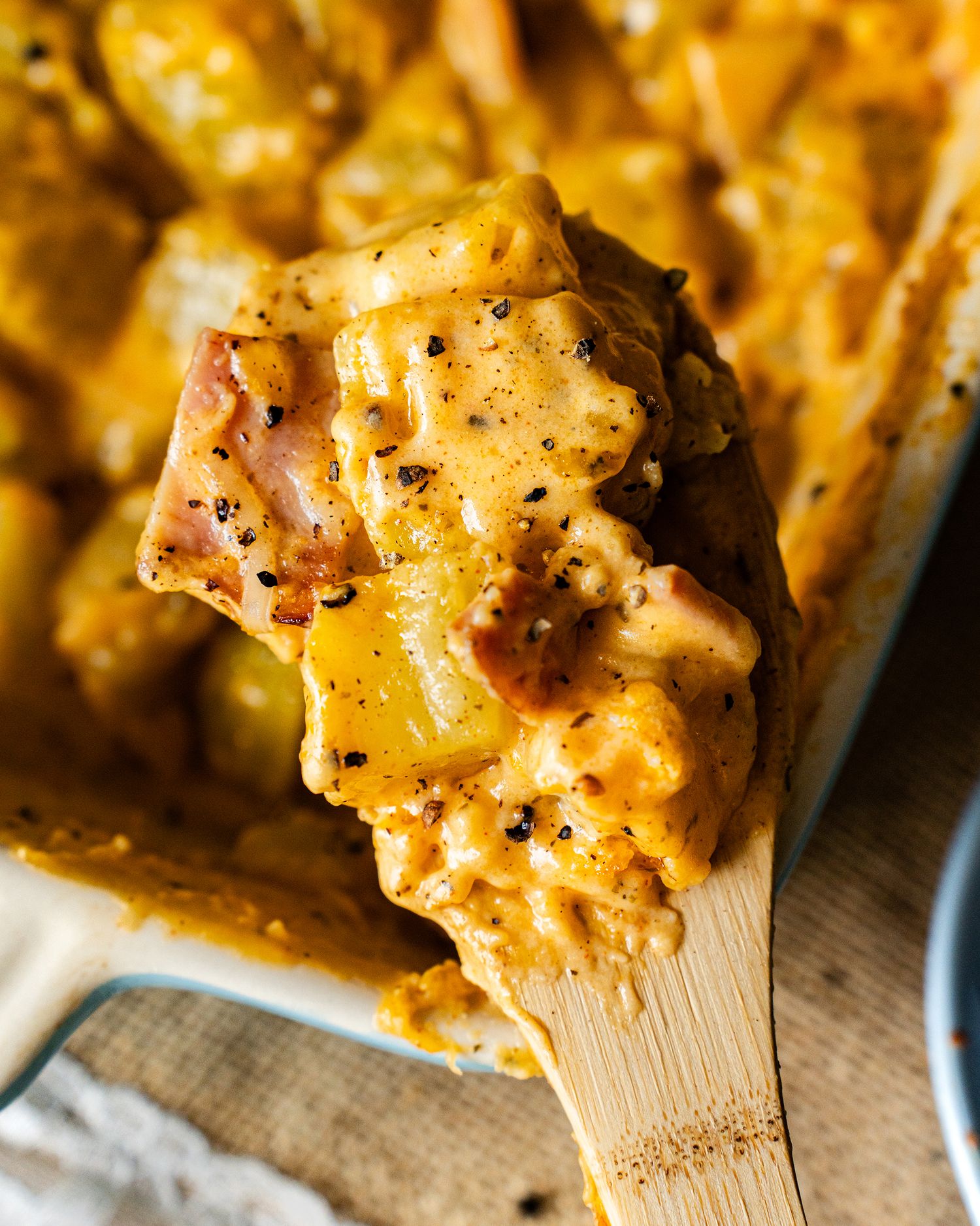 Close up of cheesy ham and potato casserole being scooped with a wooden spoon showing creamy melted cheese texture.