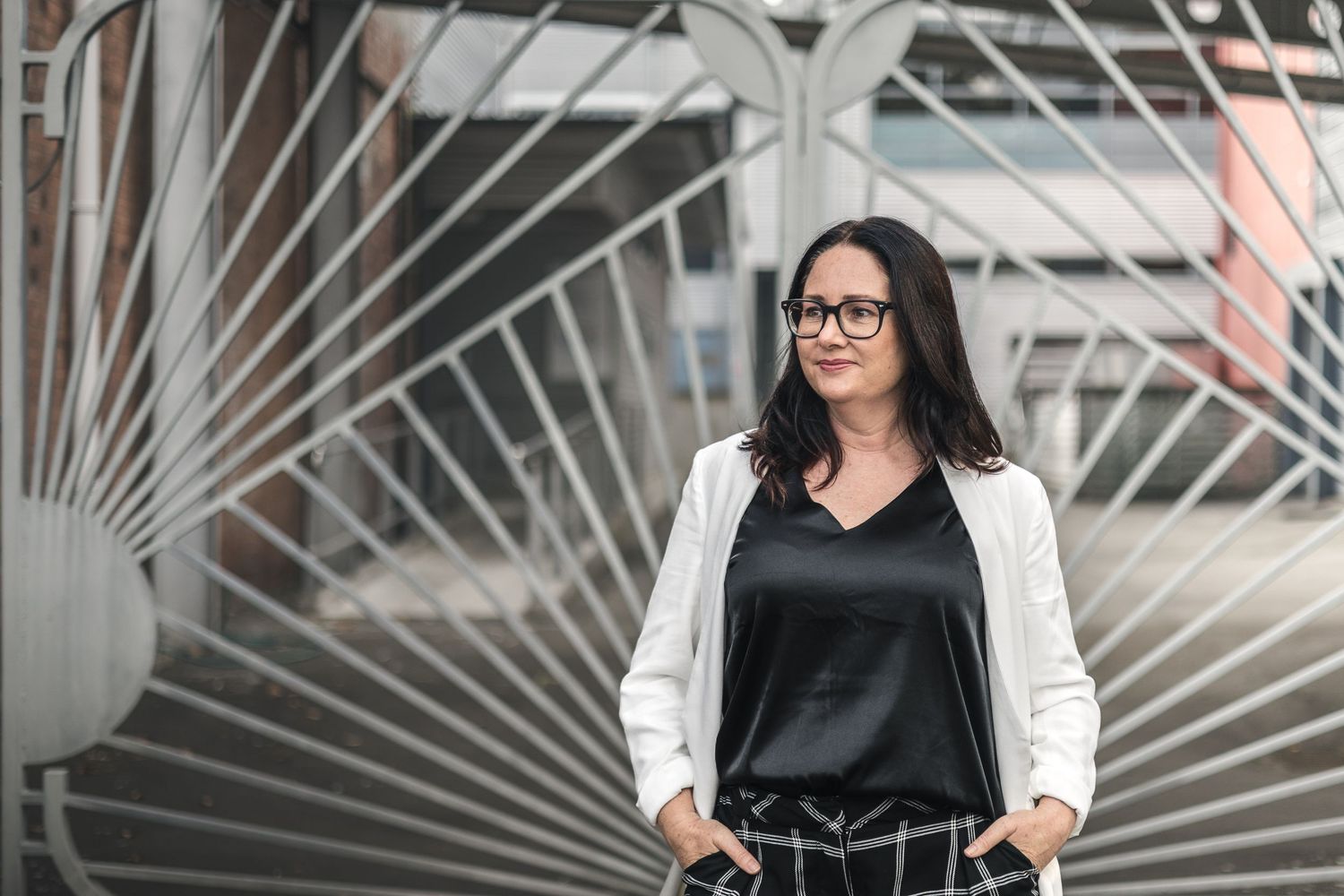 Woman in white blazer and black top poses confidently outdoors near modern geometric metal structure.