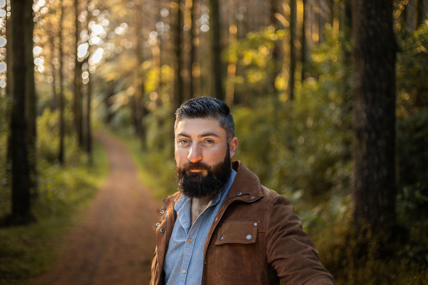 Someone with a dark beard wearing a brown jacket stands on a forest path during golden hour.