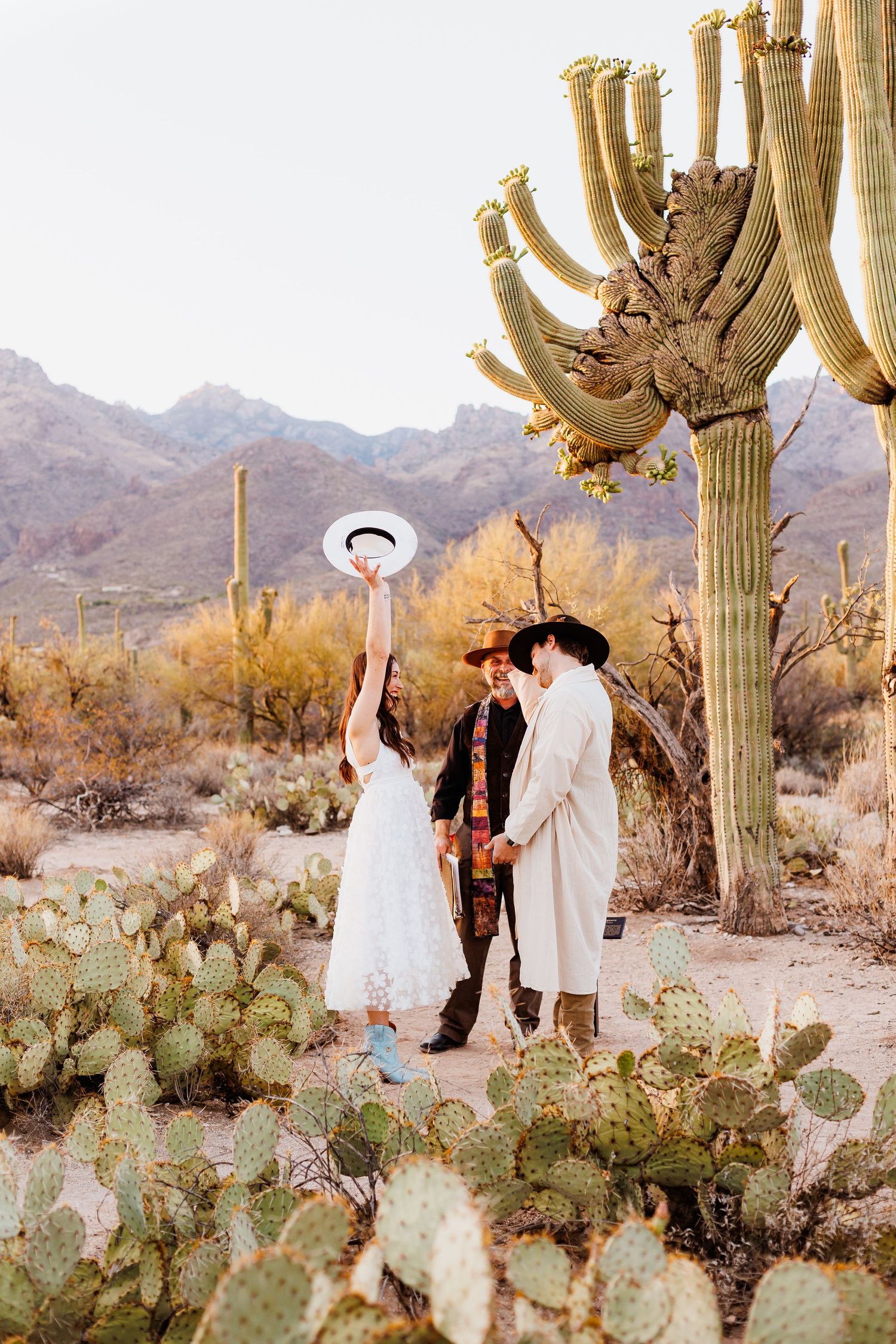 elopement Couple in white outfits and cowboy hats married beneath towering saguaro cactus in sabino canyon in Tucson arizona at sunset.