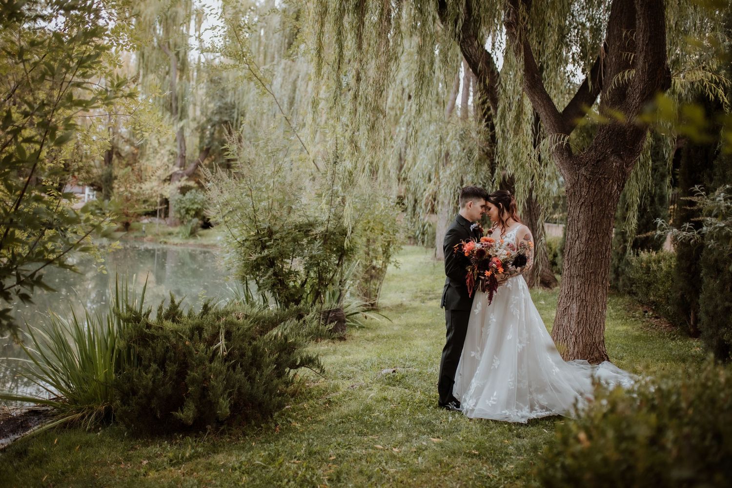 A couple in wedding attire embraces beneath weeping willow trees in an outdoor garden setting during daytime by willcox wedding photographer